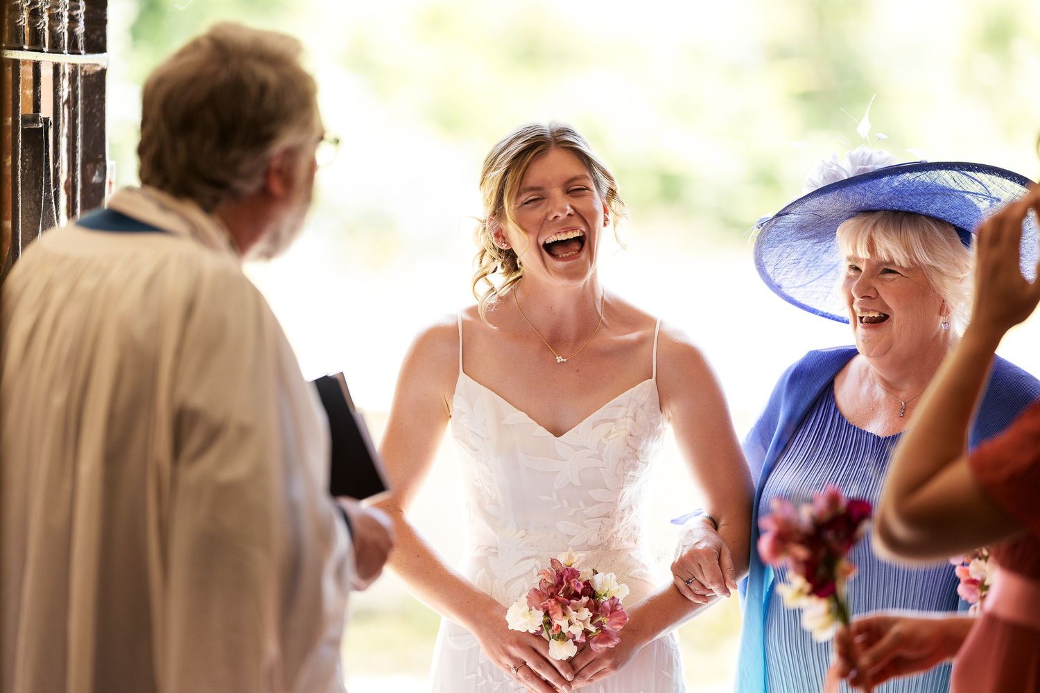 Joyful bride laughs with officiant and guest in blue hat at church wedding.