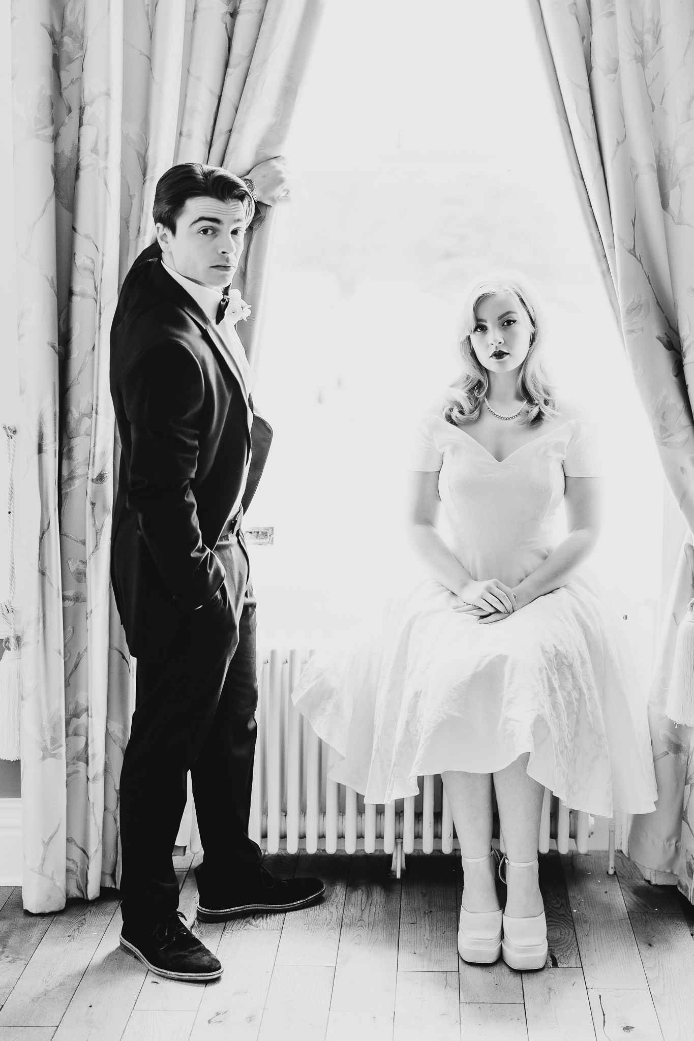 Couple in vintage attire pose dramatically by window in black and white photo.
