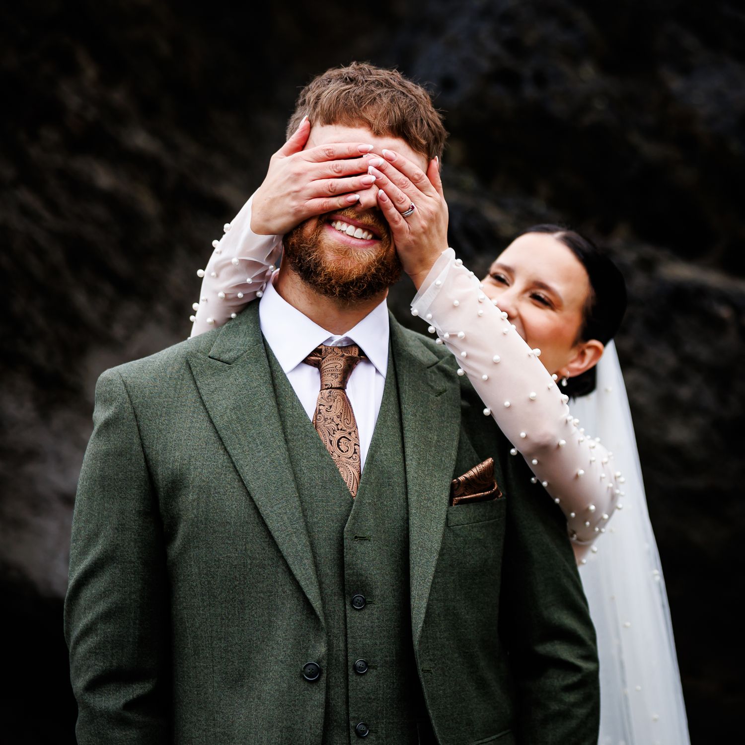 Playful bride covers her groom's eyes from behind, both wearing elegant wedding attire in an outdoor setting.