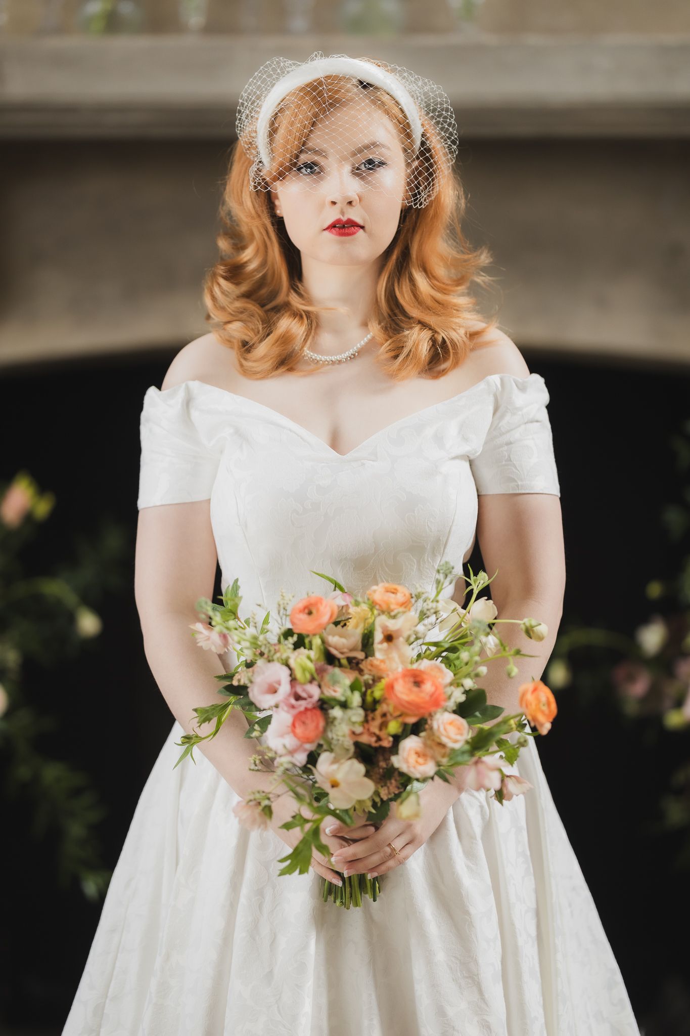 Bride in vintage style tea length dress with off-shoulder neckline holding peach and pink bouquet.