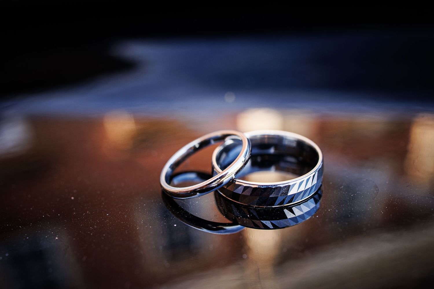 Close-up of two wedding rings with detailed metalwork on reflective surface.