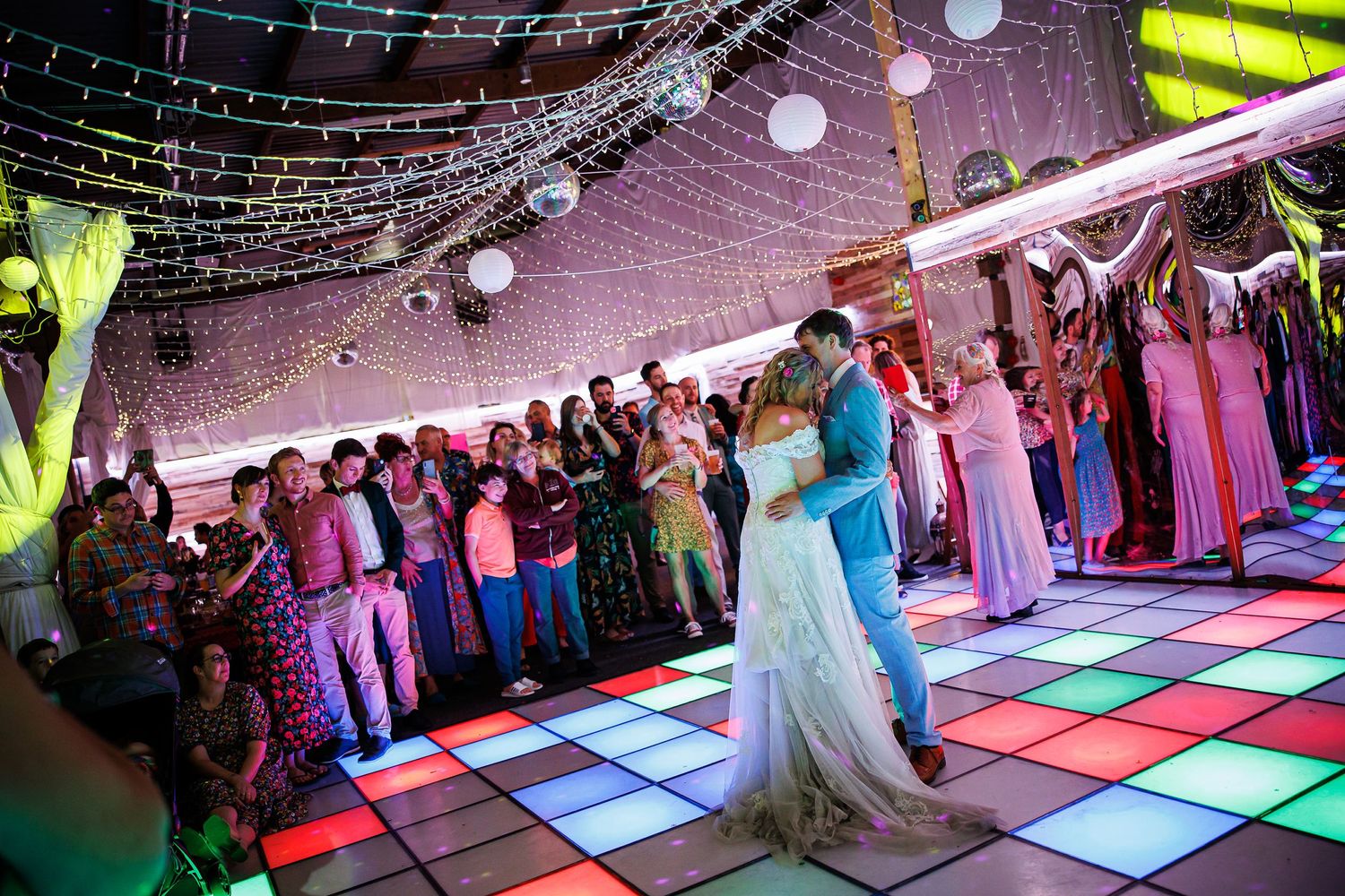 Wedding celebration with crowd surrounding couple on colorful light-up dance floor.