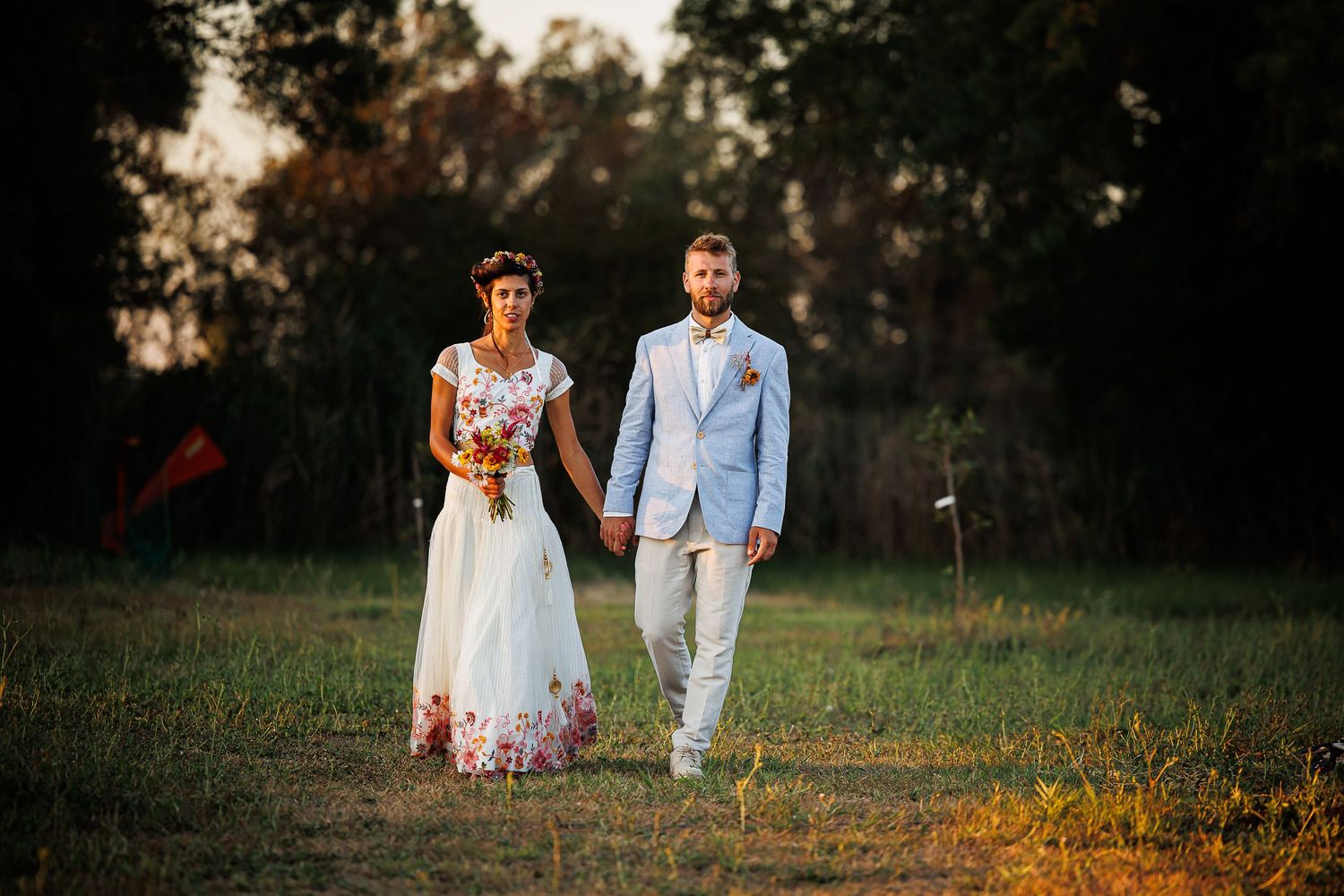 A couple walks hand in hand through a sunlit meadow at sunset wearing a white dress and light blue suit.