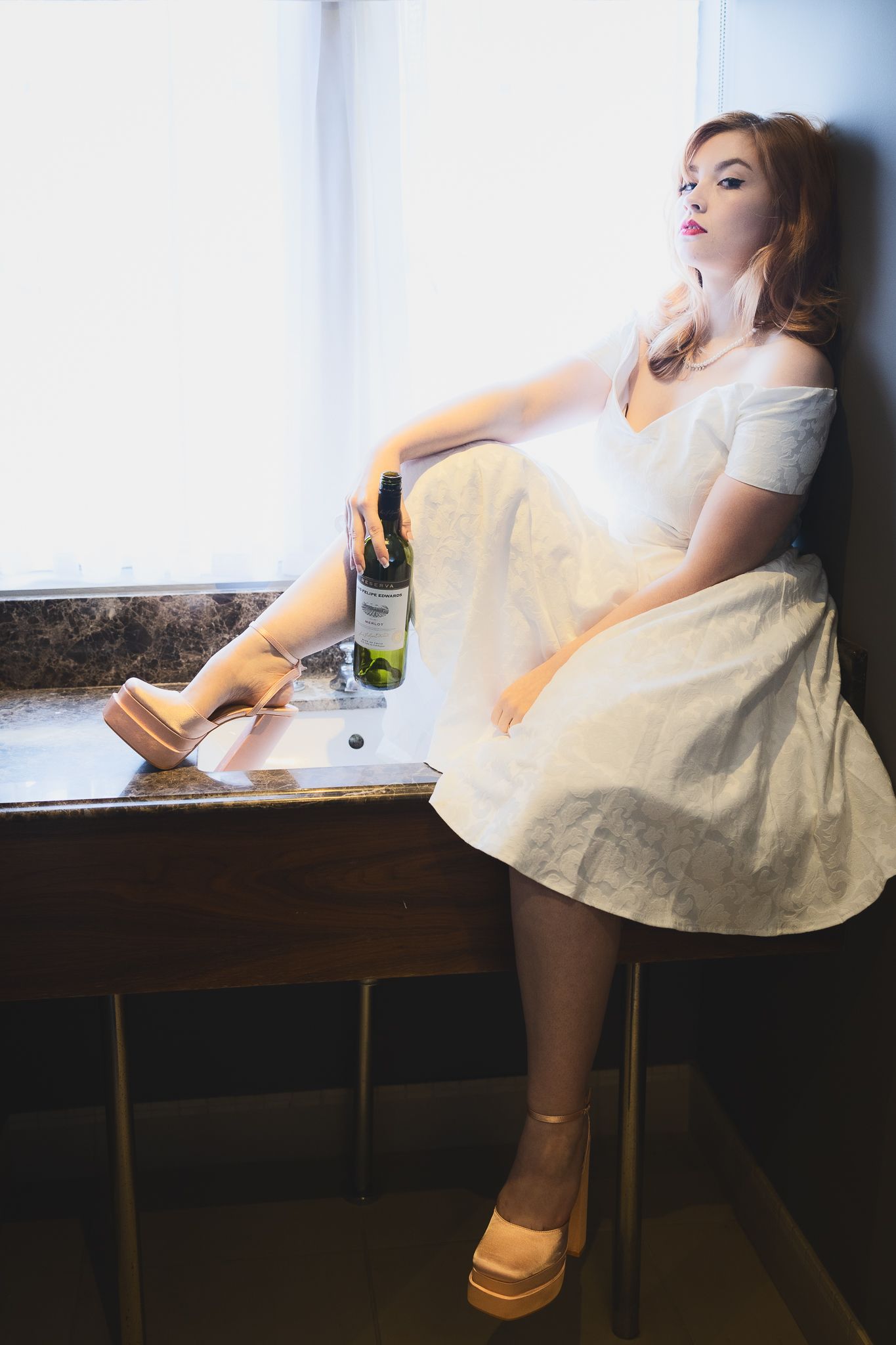 Retro styled bride in white dress and platform shoes sits near window with wine bottle.
