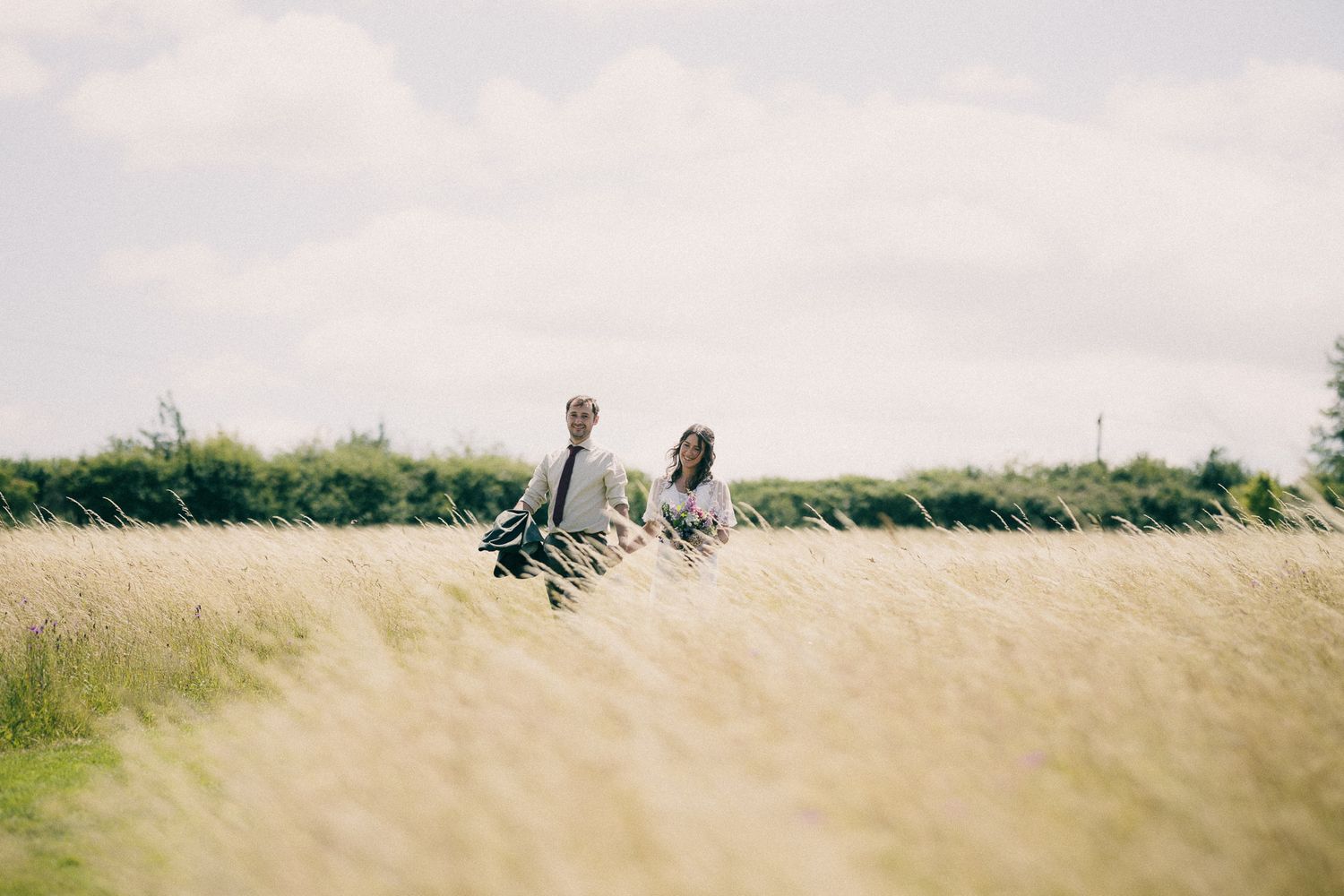 A bride and groom smile at each other in a sunny field, she holds a wildflower bouquet as he carries his jacket over his shoulder.