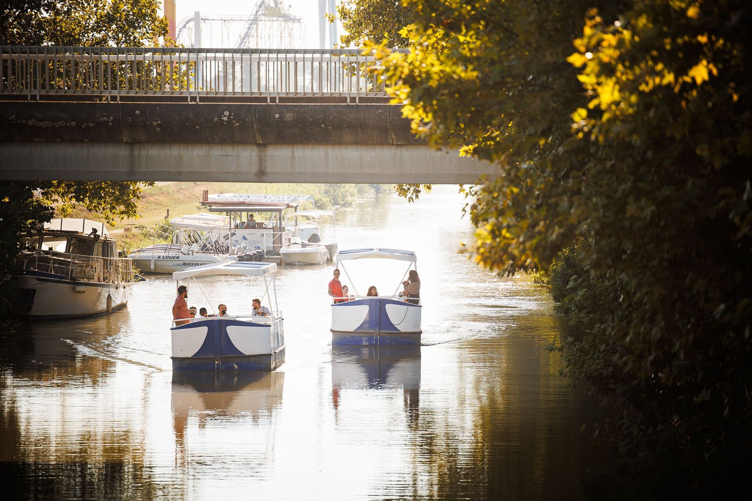 Small boats glide under a bridge on calm waters during a sunny afternoon.