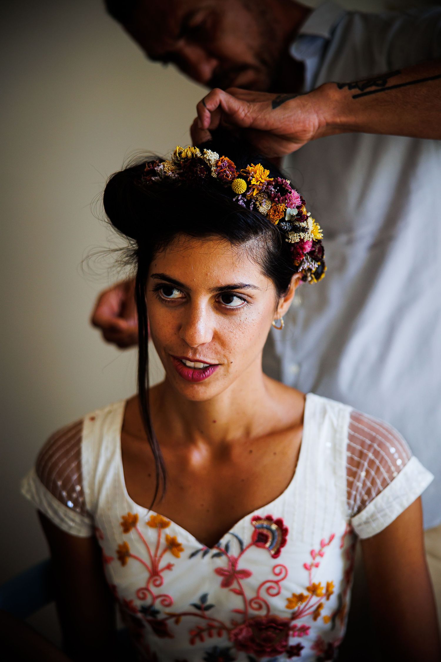 A woman in a floral dress getting flowers arranged in her hair during wedding preparation.