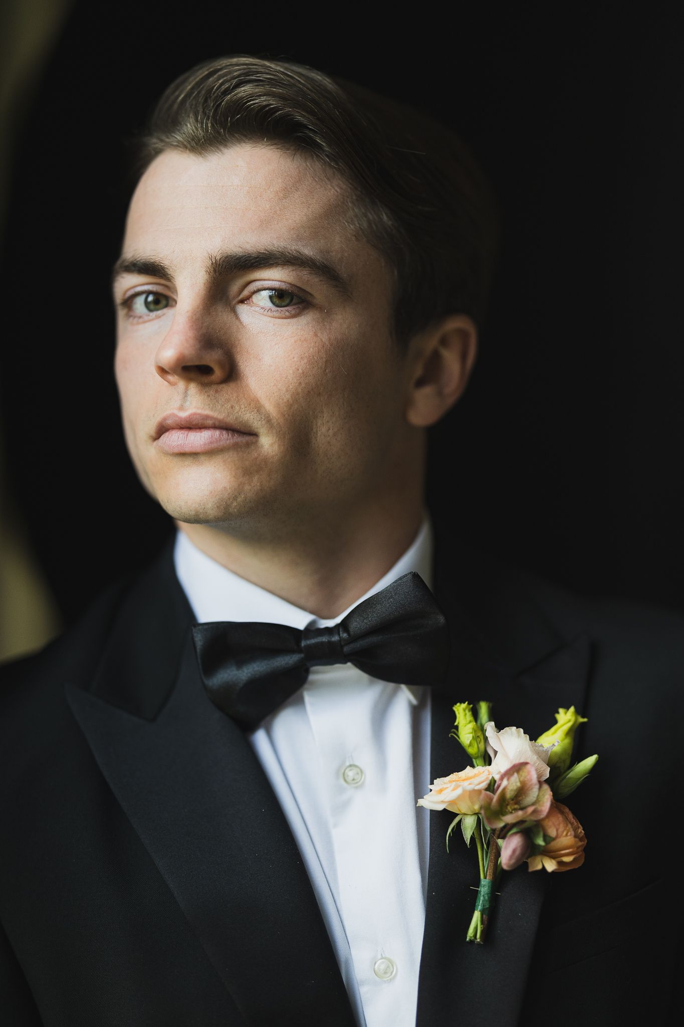 Portrait of groom in black tuxedo with peach floral boutonniere against dark background.