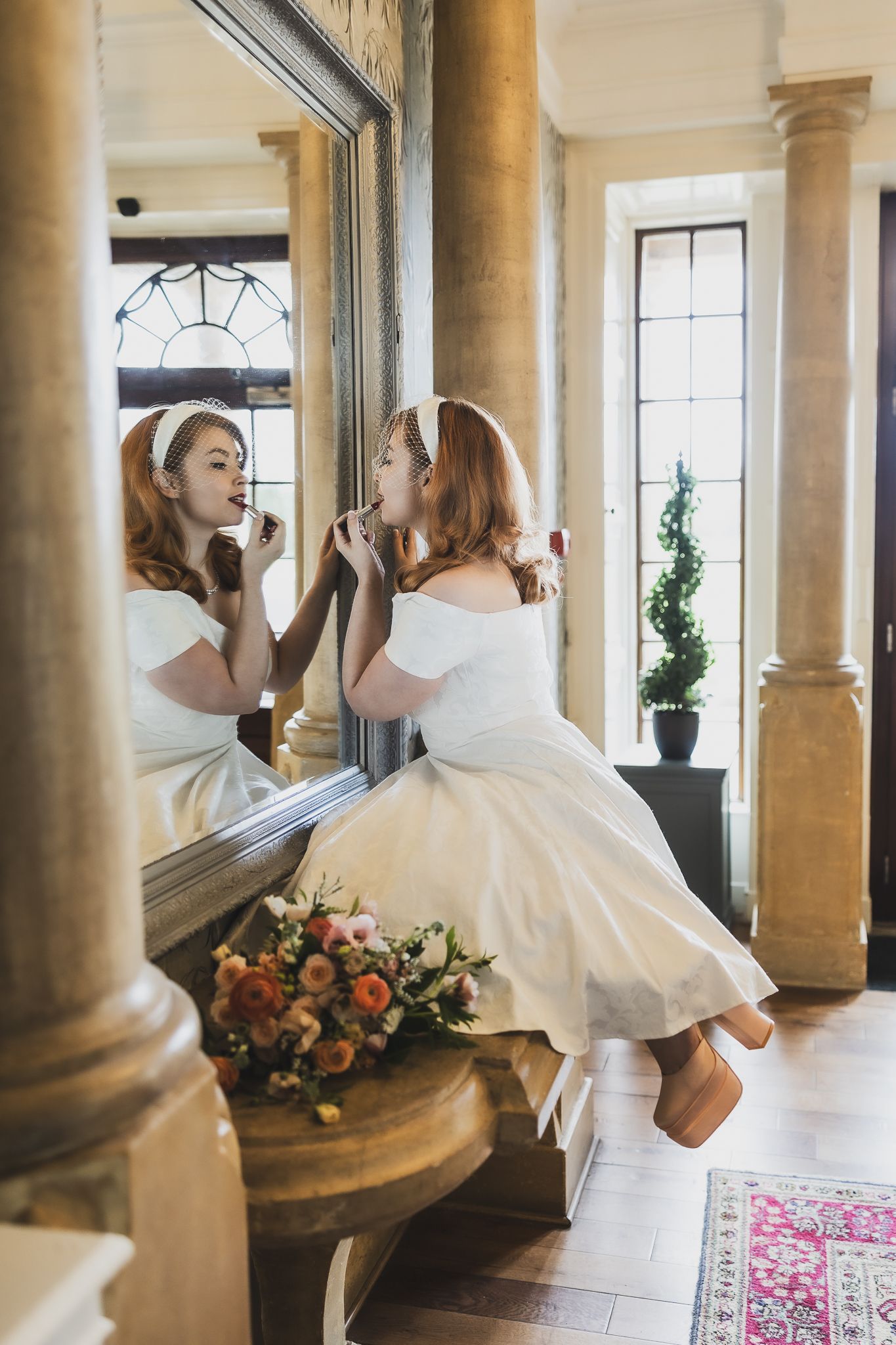Whimsical portrait of bride applying lipstick in ornate mirror with classical architecture.