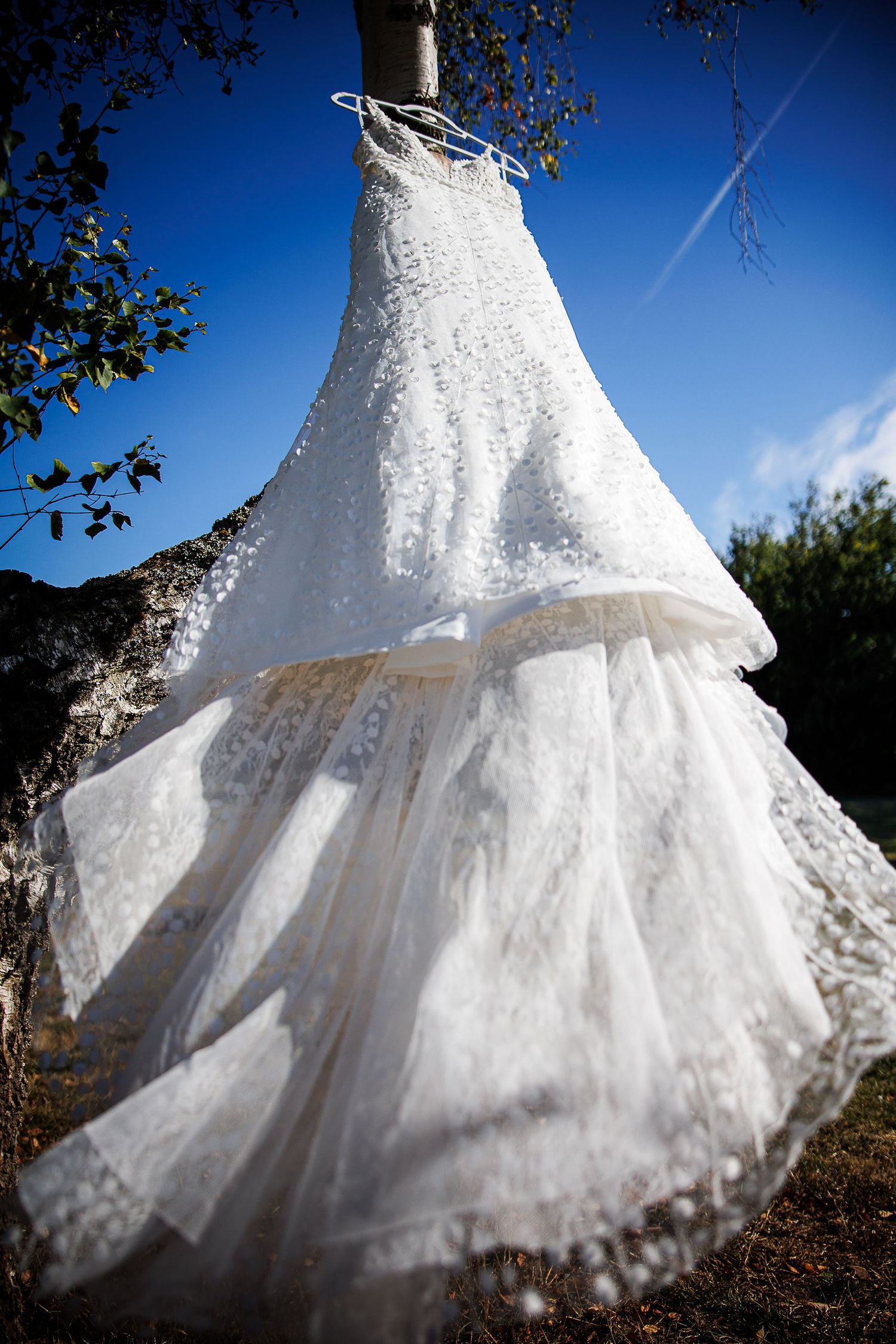 An elegant lace wedding dress hanging outdoors against bright blue sky.