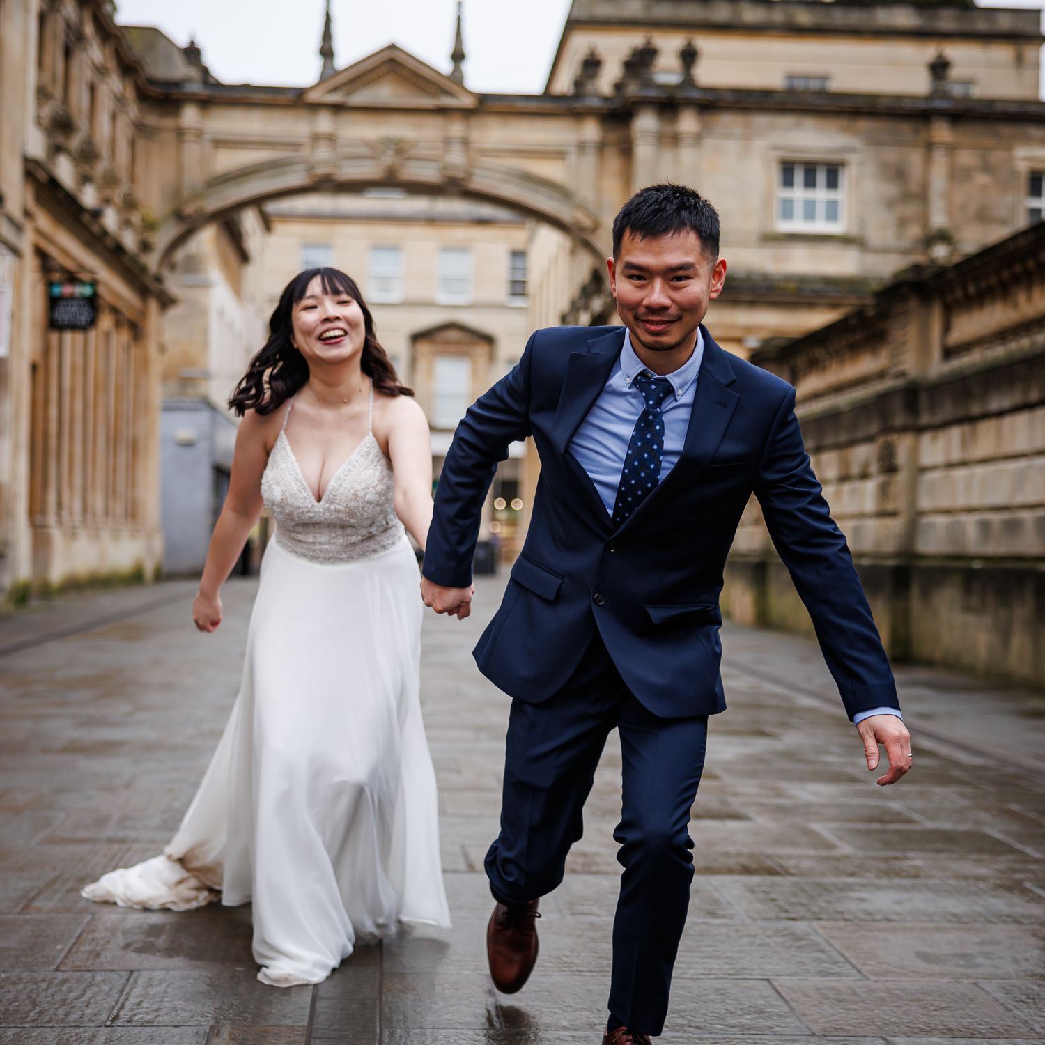 A joyful couple in wedding attire runs together down a historic stone walkway with elegant architecture in the background.