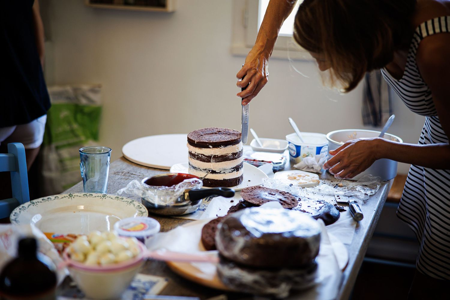 Someone decorating layered chocolate cakes on a table with various baking supplies.