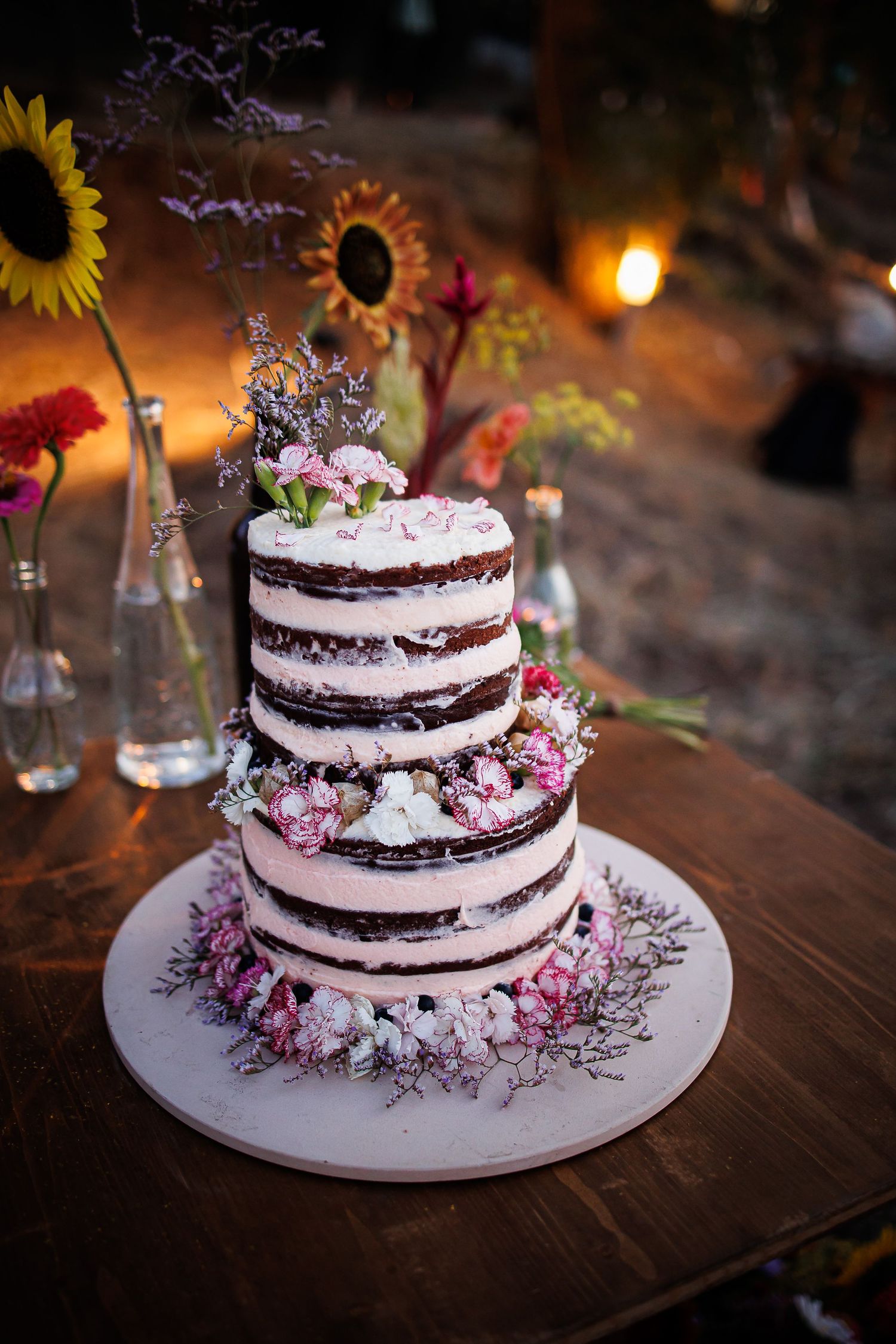 A rustic semi-naked wedding cake decorated with pink flowers and sunflowers on a wooden table.