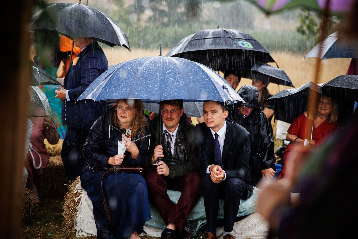 Wedding guests huddle together under black umbrellas during a rainy outdoor ceremony.