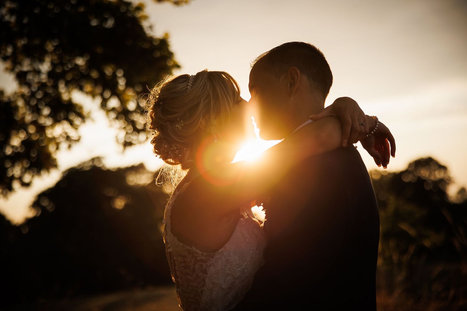 A silhouetted couple embraces during golden hour with sun rays peeking through trees in the background.