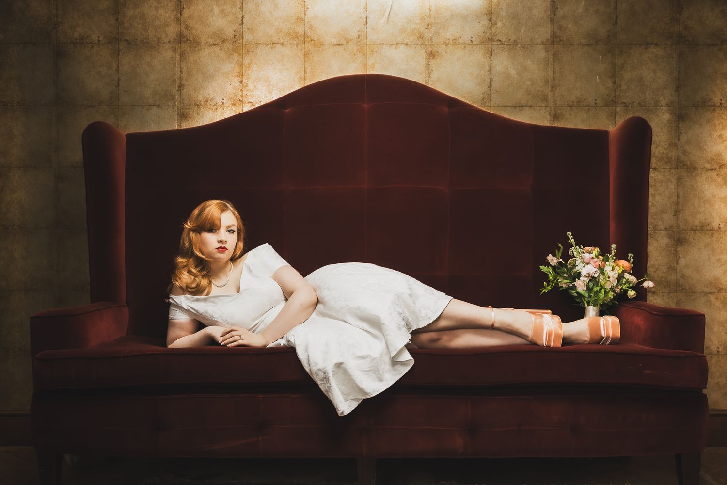 Romantic vintage-inspired bridal portrait on velvet sofa with floral arrangement.