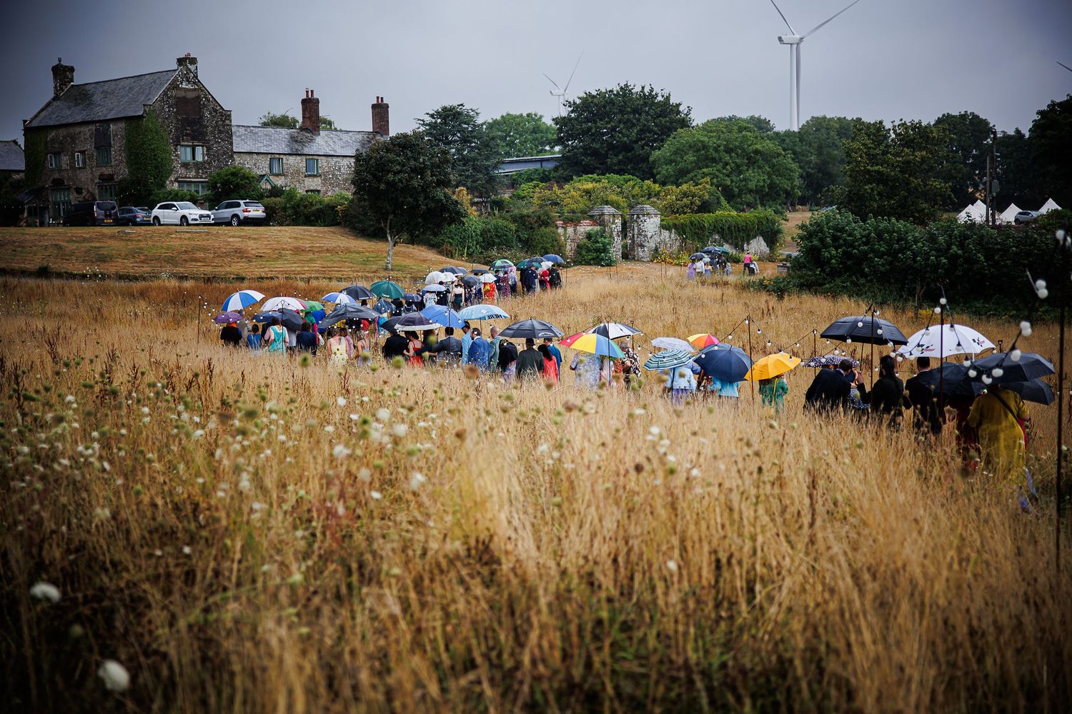 Blue umbrellas dot a golden wheat field as wedding guests gather for an outdoor ceremony.