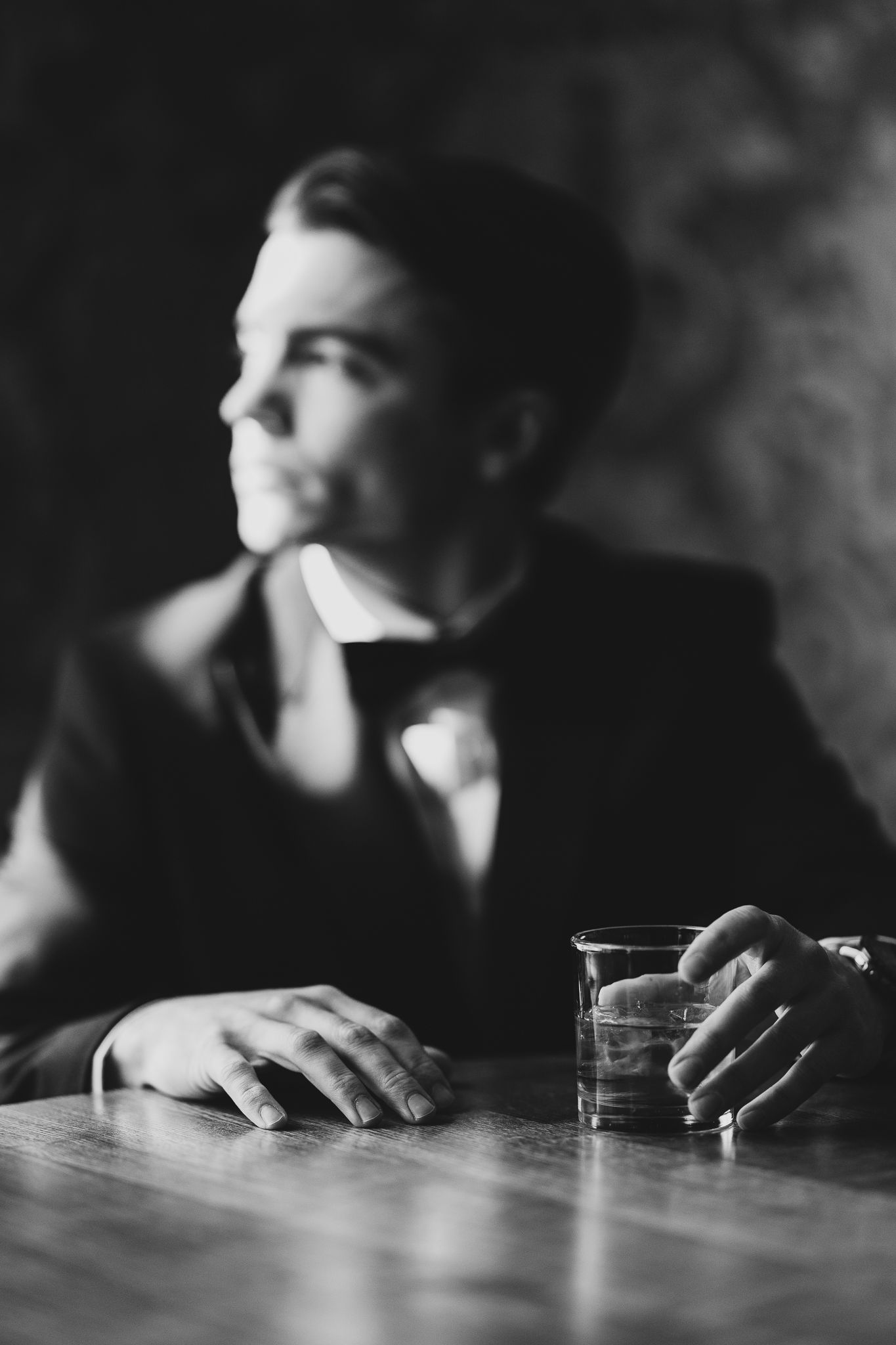 Black and white photo of groom in tuxedo sitting at table with drink in vintage style portrait.