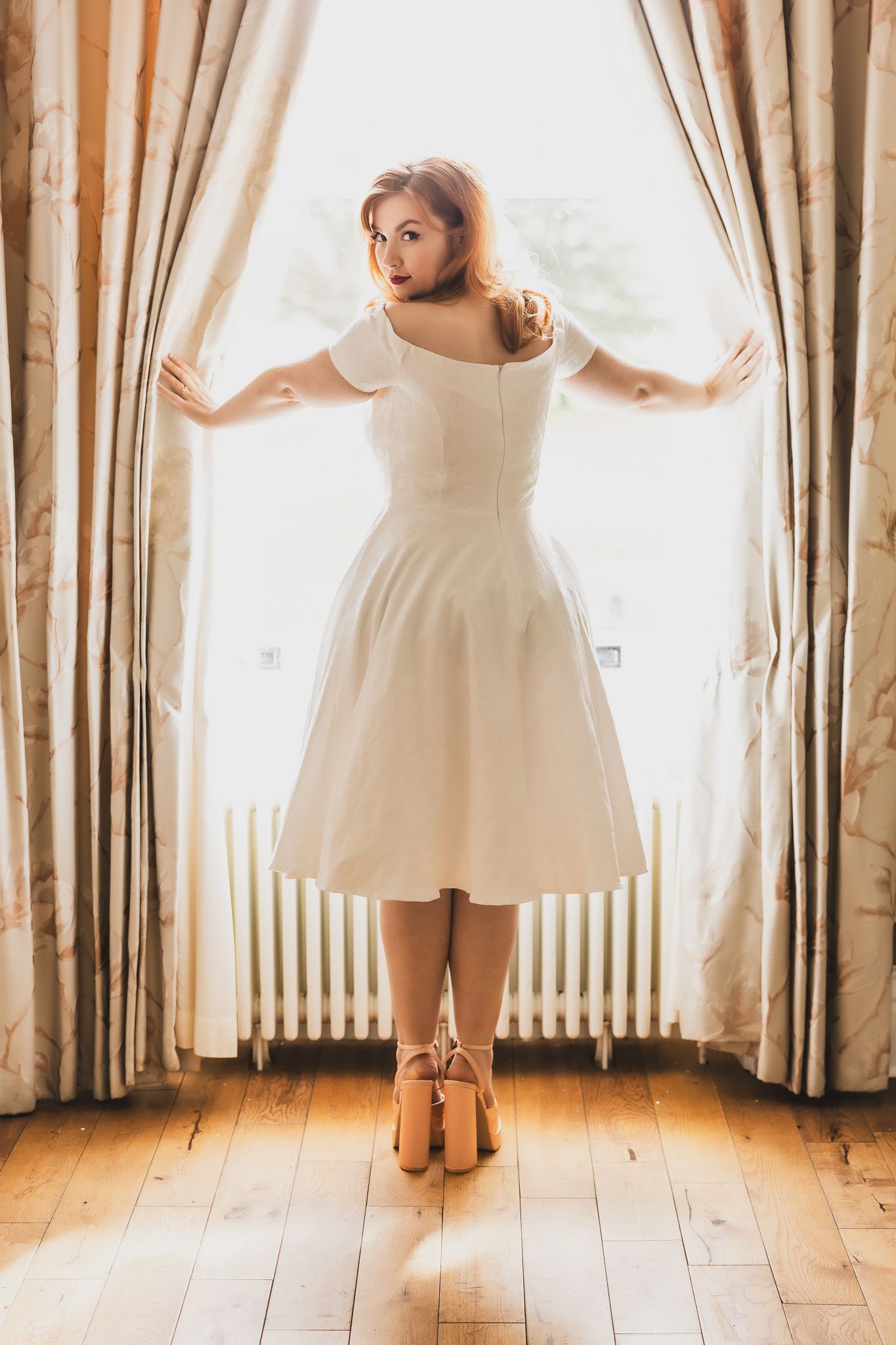 Elegant bride poses between curtains in sunlit room.