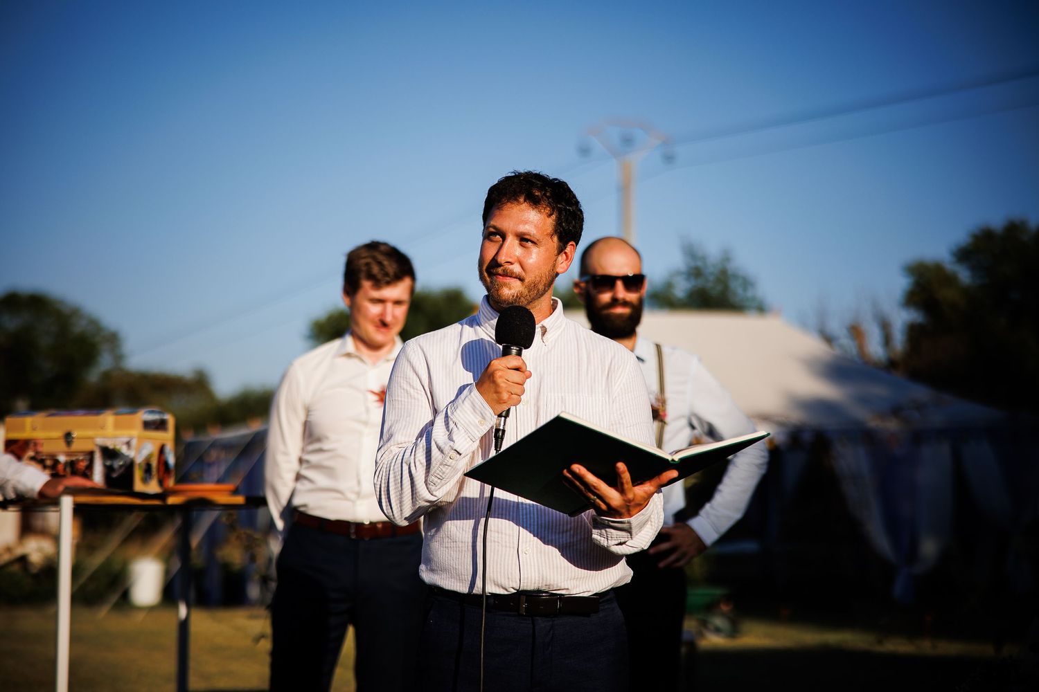 Speaker holds microphone and book during outdoor ceremony.