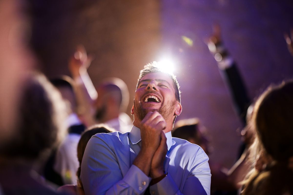 Excited man clapping and laughing at a concert, bathed in bright stage light among a cheering crowd.