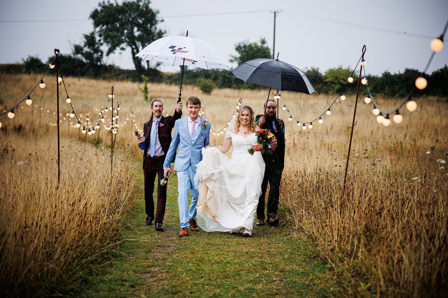 Wedding party walks through tall grass field with string lights and umbrellas.