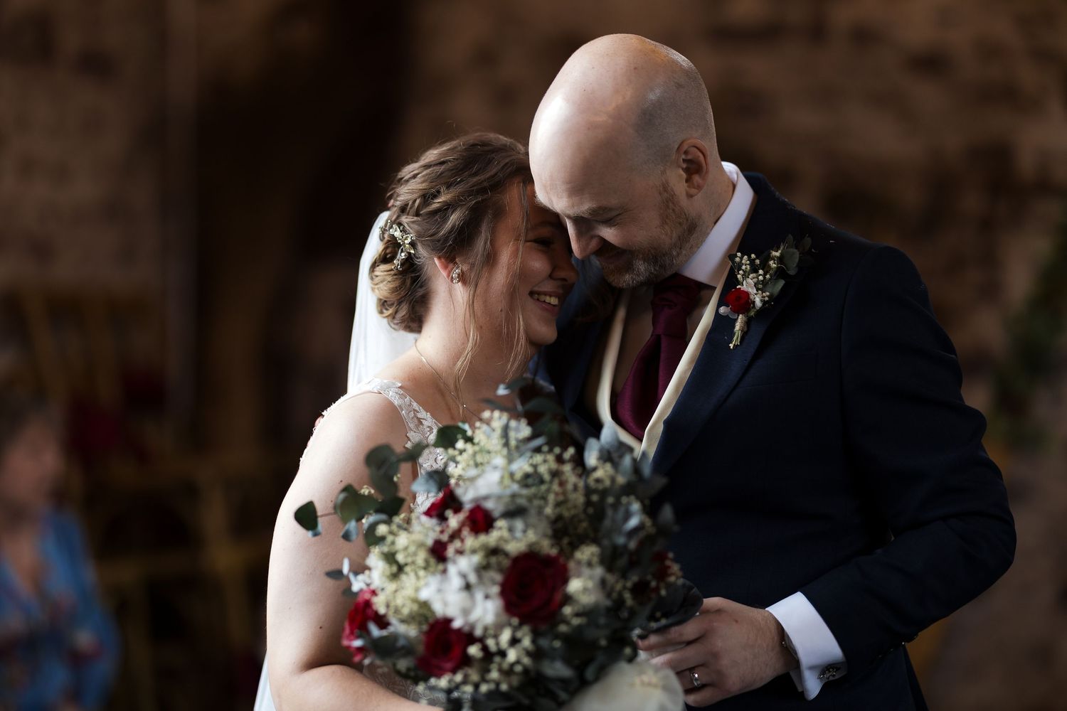 Bald groom tenderly embraces smiling bride holding red and white bouquet at wedding.