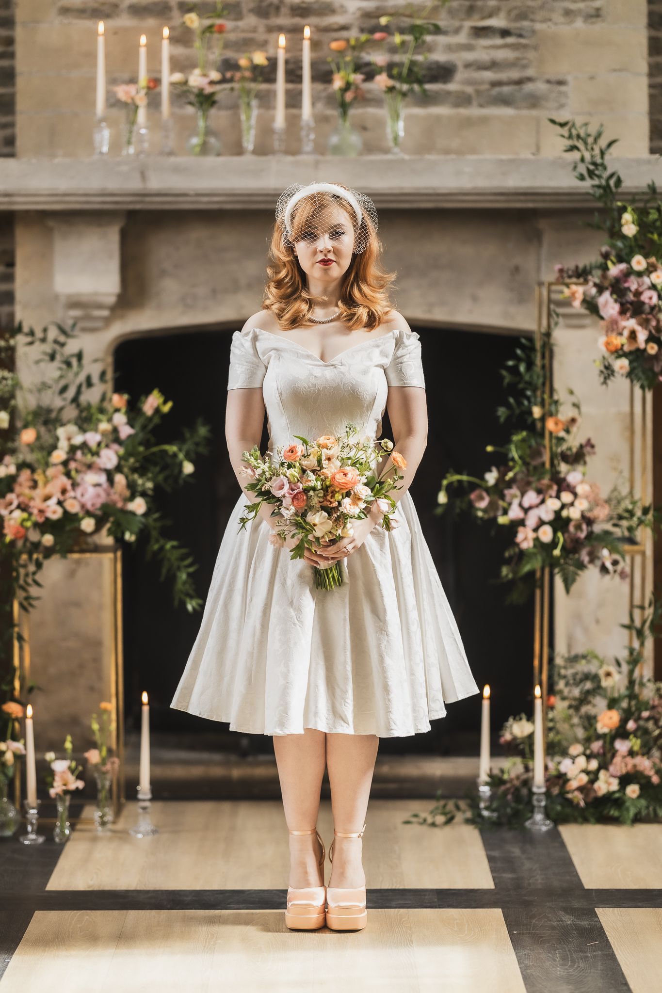 Bride in 1950s style tea length dress stands before fireplace decorated with candles and flowers.