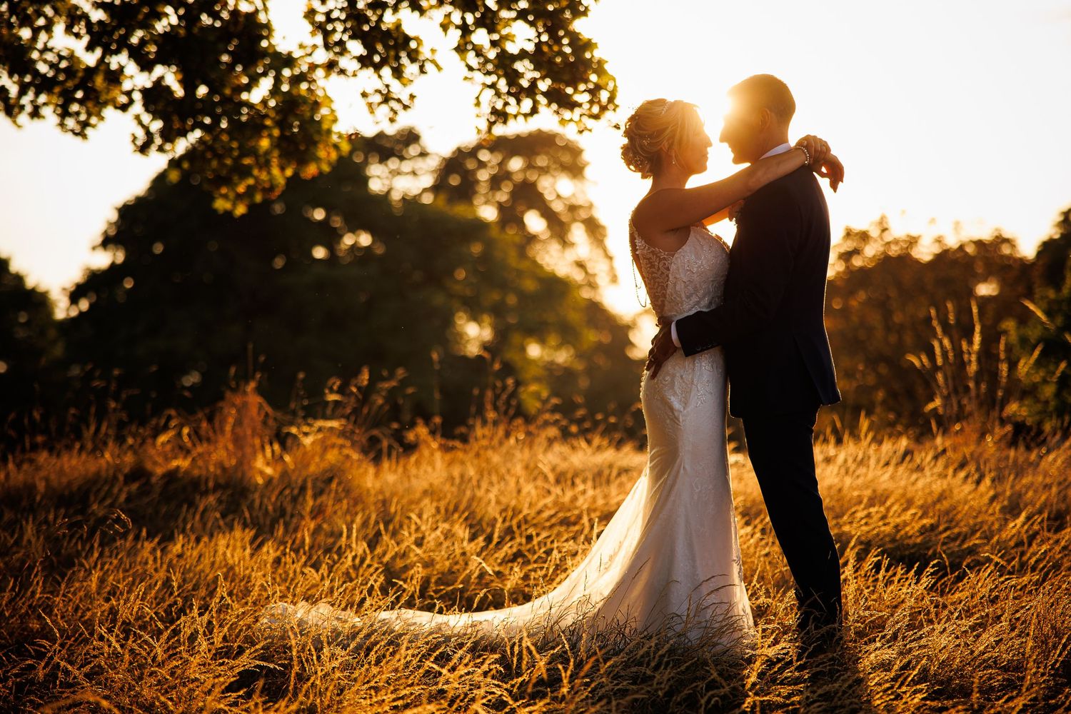 A romantic silhouette of a couple in wedding attire embracing in a sunlit golden field at sunset.