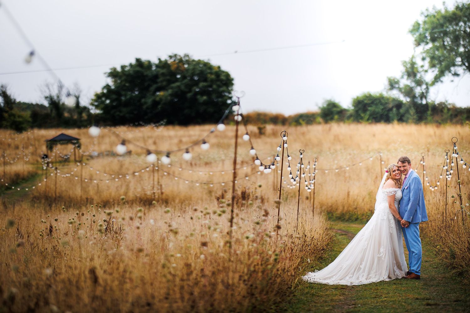 Newlyweds in light blue and white stand in golden field during sunset.