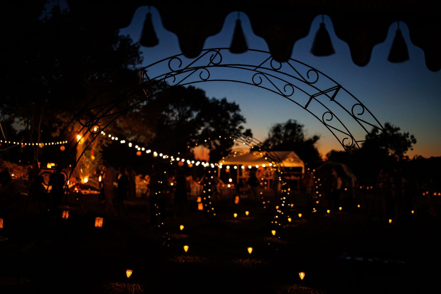 String lights illuminate an outdoor wedding venue at dusk under a decorative arch.