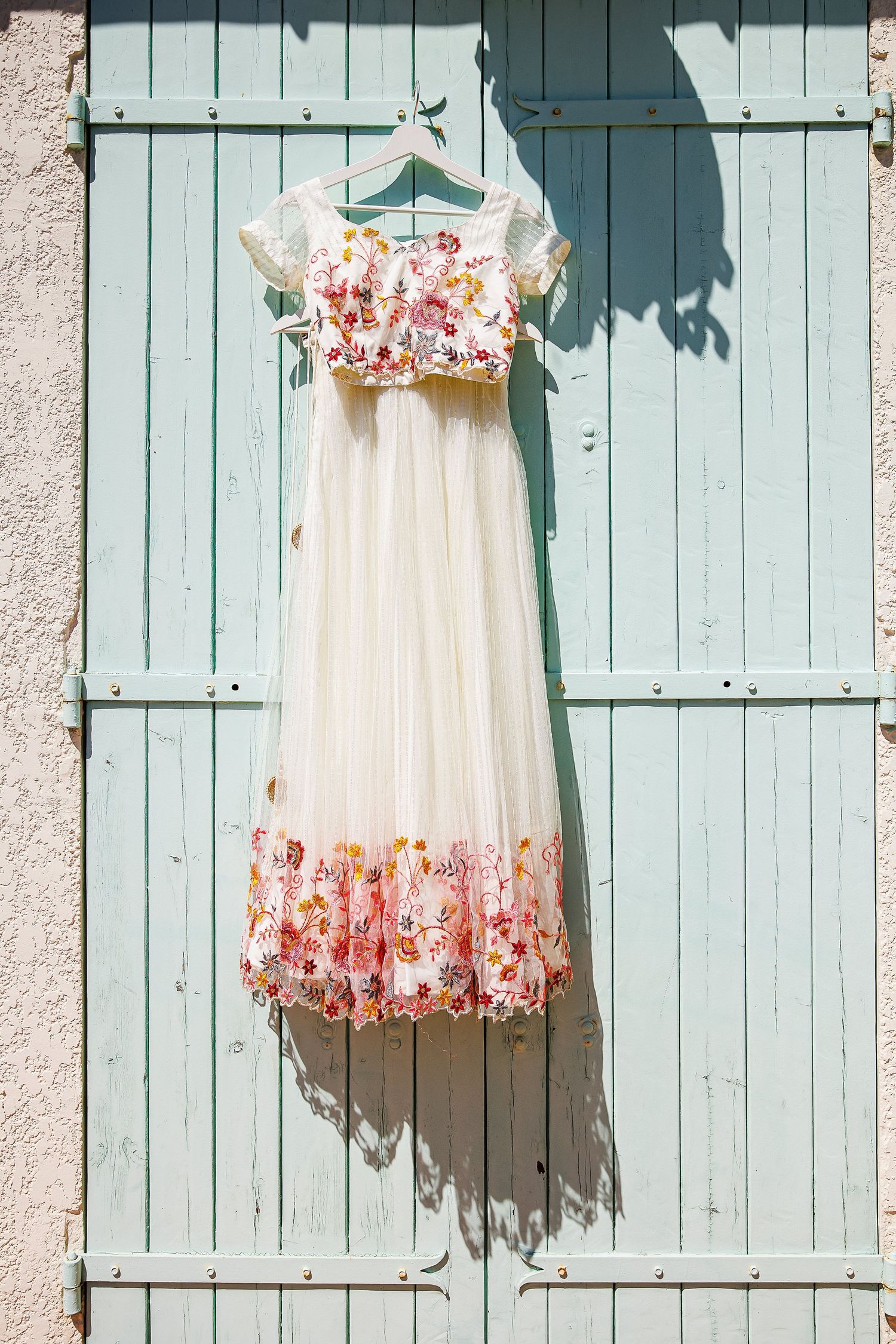 A delicate white dress with floral details hanging on a blue-green shuttered wall.