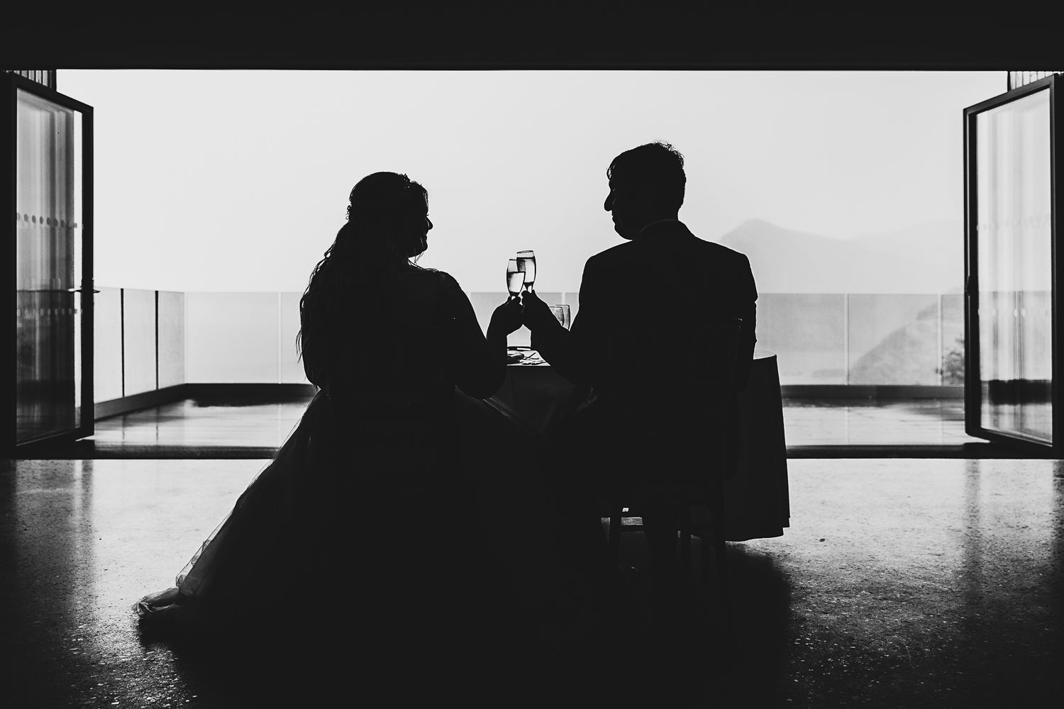 Two silhouetted figures share drinks while sitting by large windows overlooking a scenic view in black and white photography.