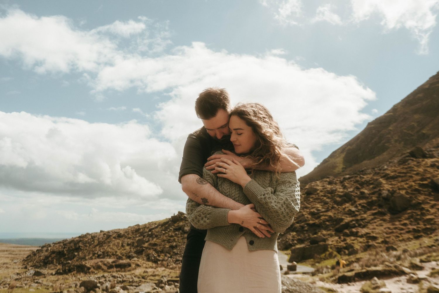 picture of couple in front of rugged green mountain, she's leaning against him and his eyes are closed