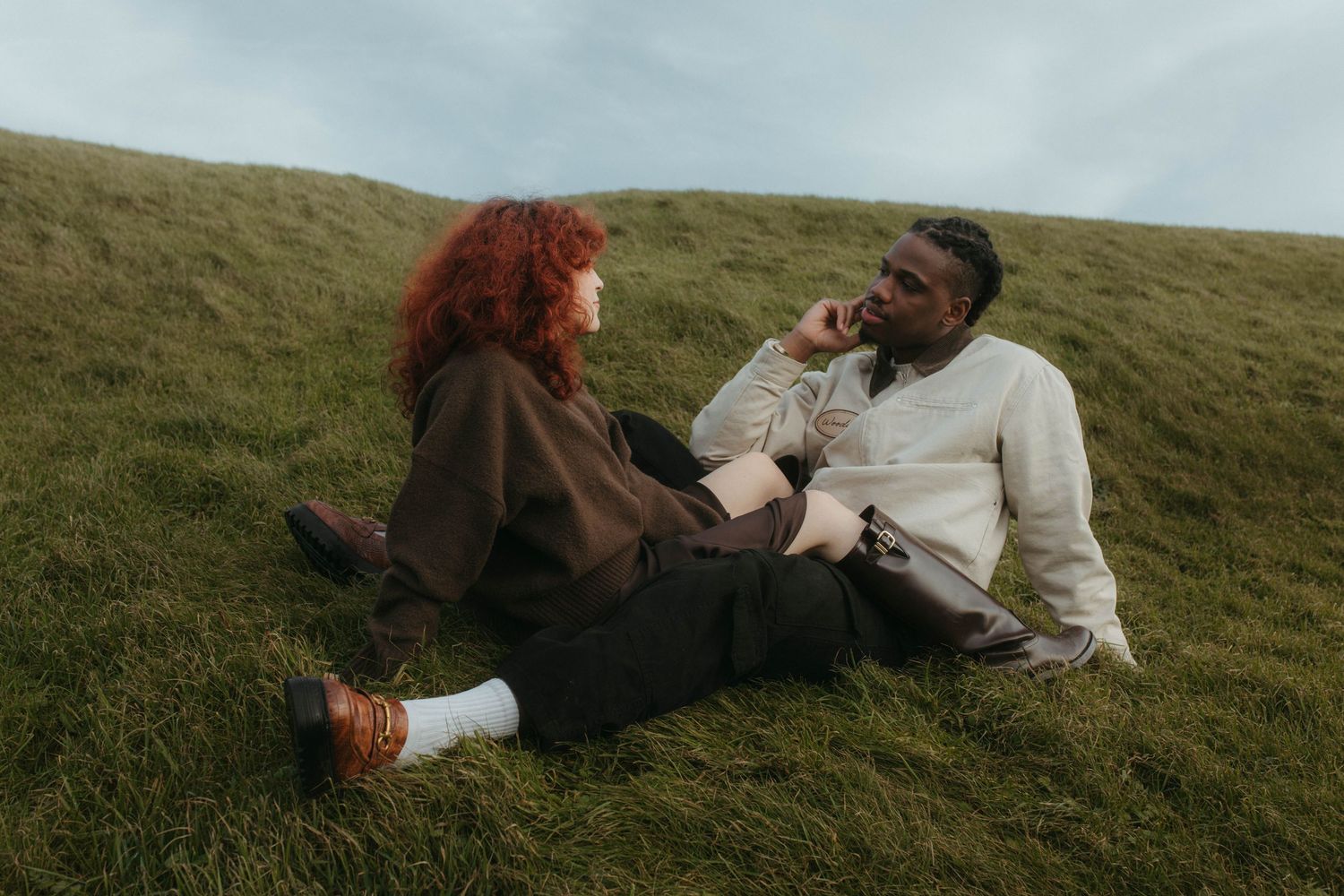 Interracial couple posing on a grassy hill, their legs overlap and they are looking at each other - couples shoot