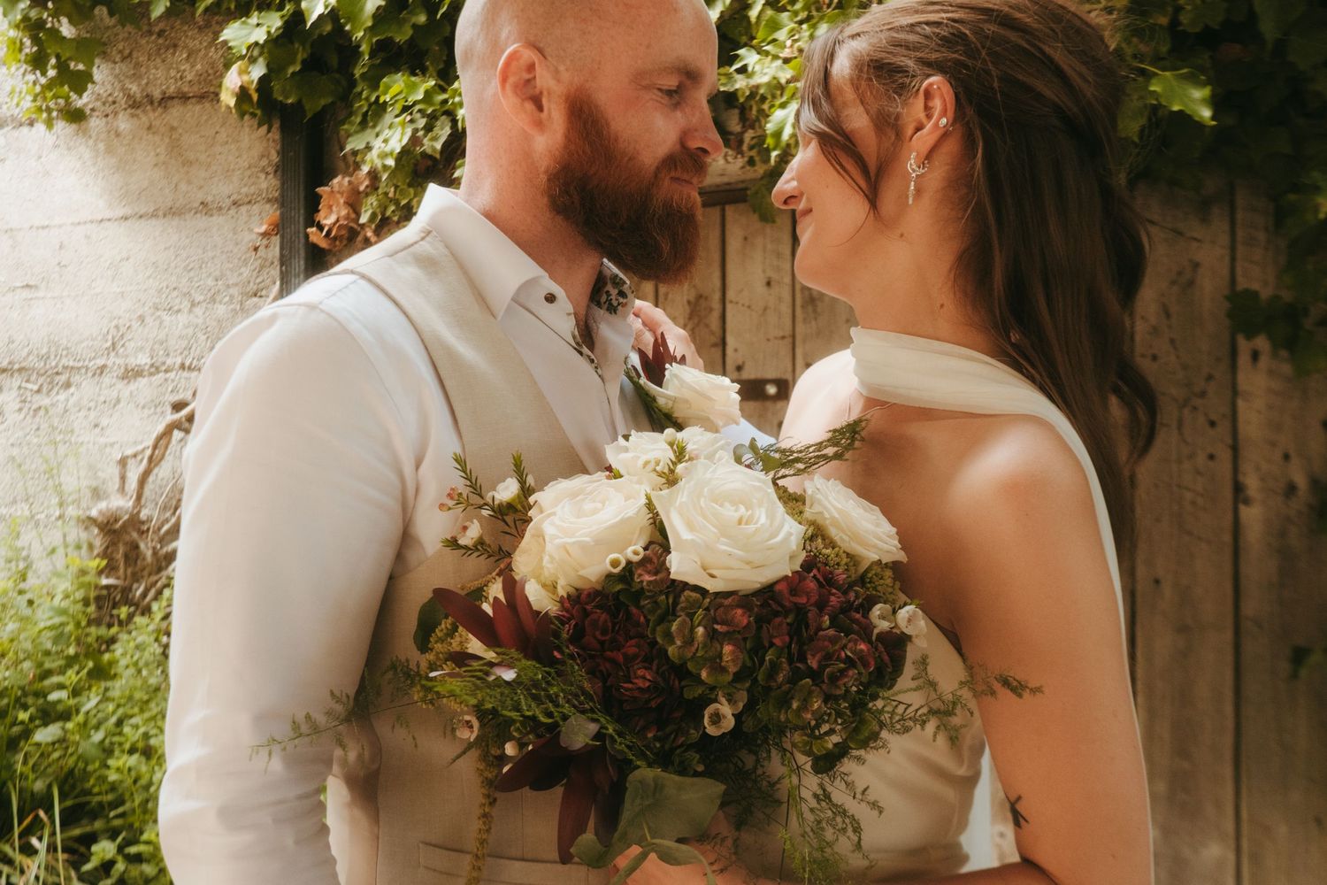 Photograph of bride and groom in a meadow after their wedding ceremony, she has dark brown hair and he is bald with a ginger beard.