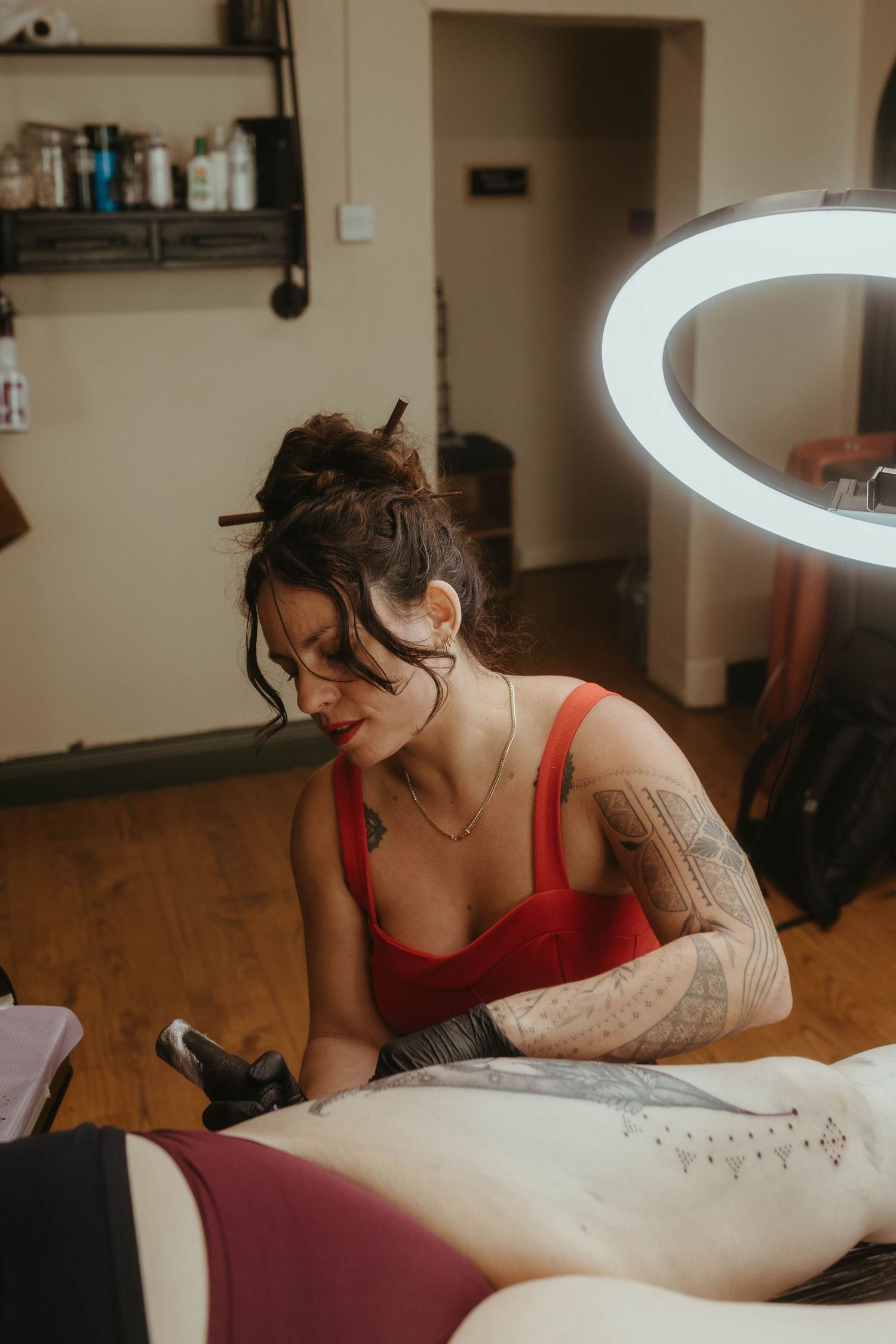 Photo of tattoo artist tattooing a client, she has her hair tied up and is wearing a red top