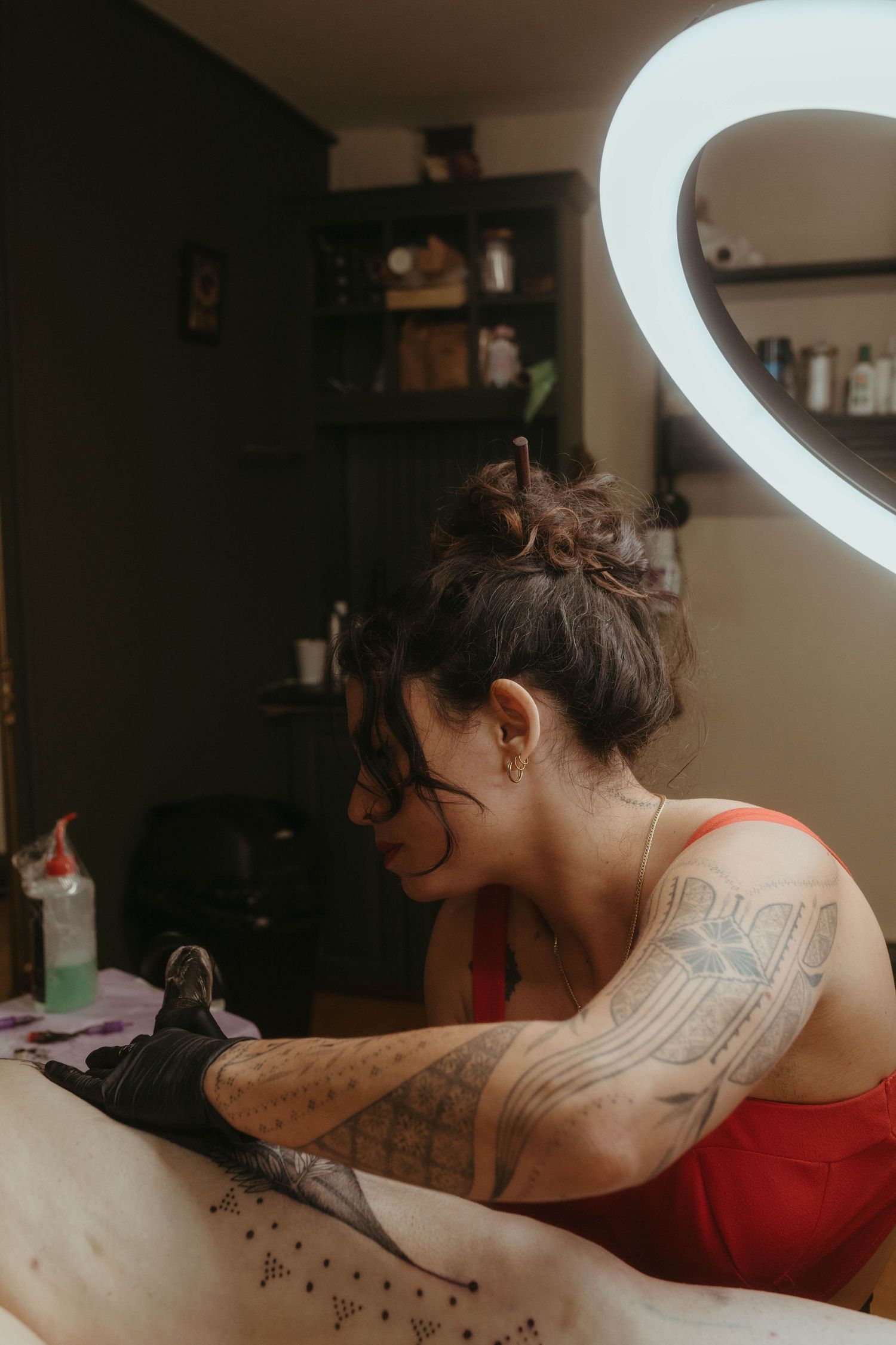 Photo of tattoo artist tattooing a client, she has her hair tied up and is wearing a red top