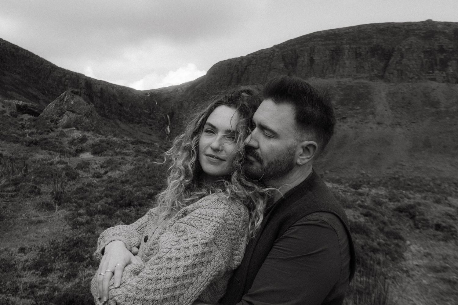 Photograph from an engagement shoot, the couple is posing in front of rugged Irish nature, he is embracing her from behind