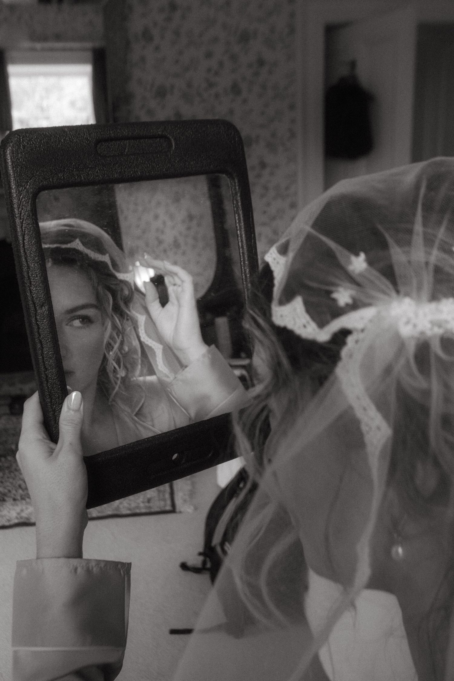 Black and white photograph of bride getting ready, focused on her looking into a mirror