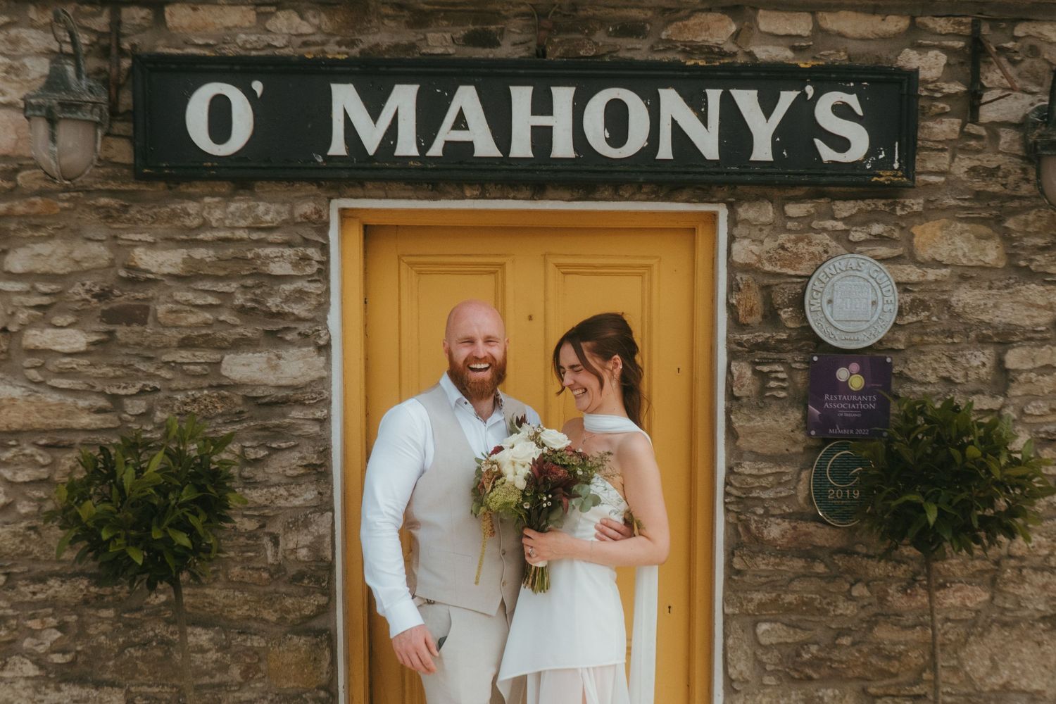 Photograph of bride and groom in a meadow after their wedding ceremony, she has dark brown hair and he is bald with a ginger beard.