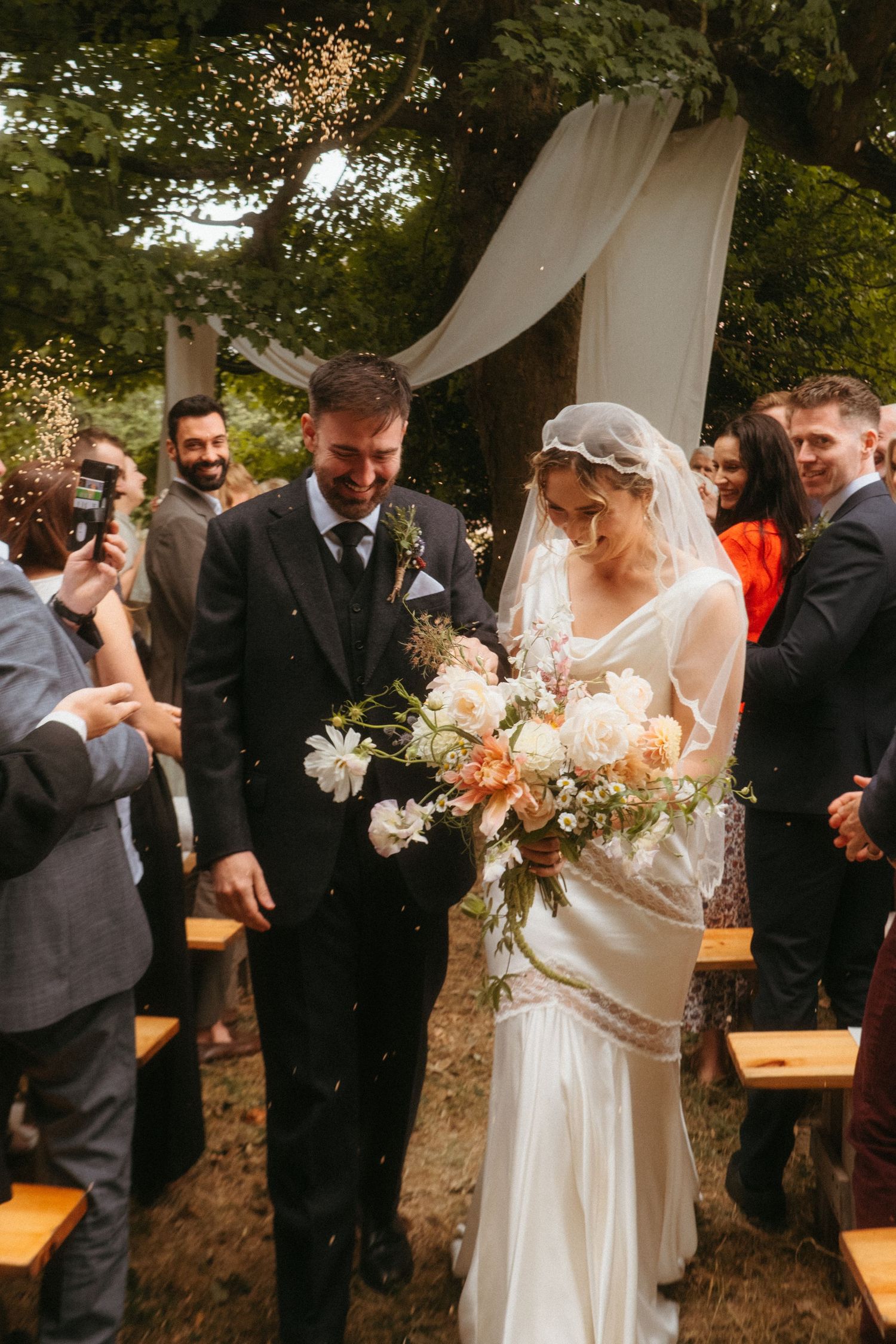 Color photograph of bride and groom exiting their wedding ceremony, barley is being thrown at them in celebration, they're smiling