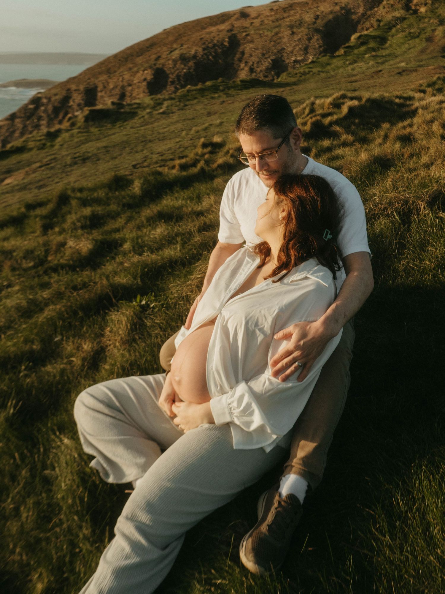 Photograph of a couple on a cliff outlooking the sea, she's leaning against him, they are sitting