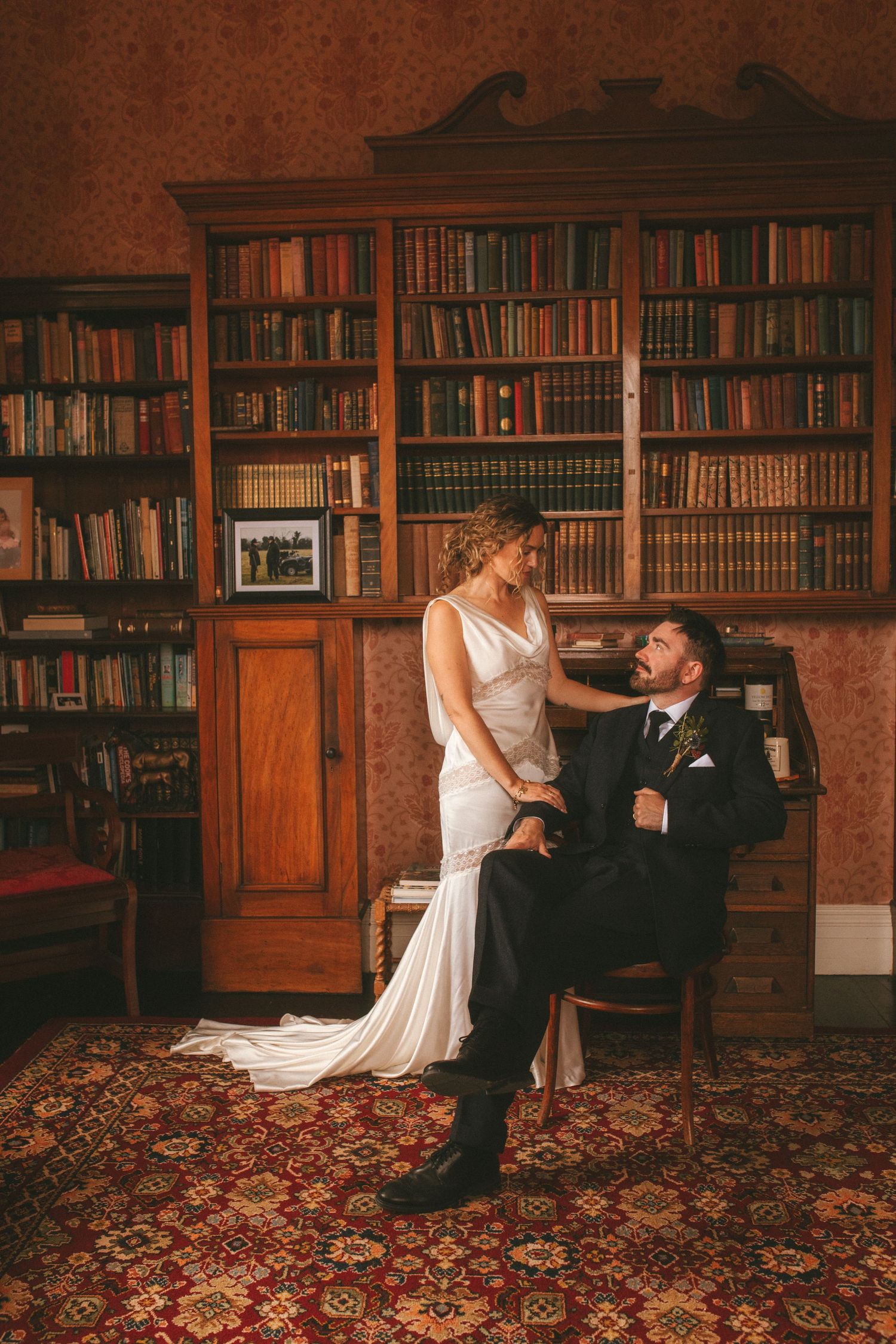 Photograph of bride and groom during their bridal session posing in an old library, he is sitting and she is standing next to him