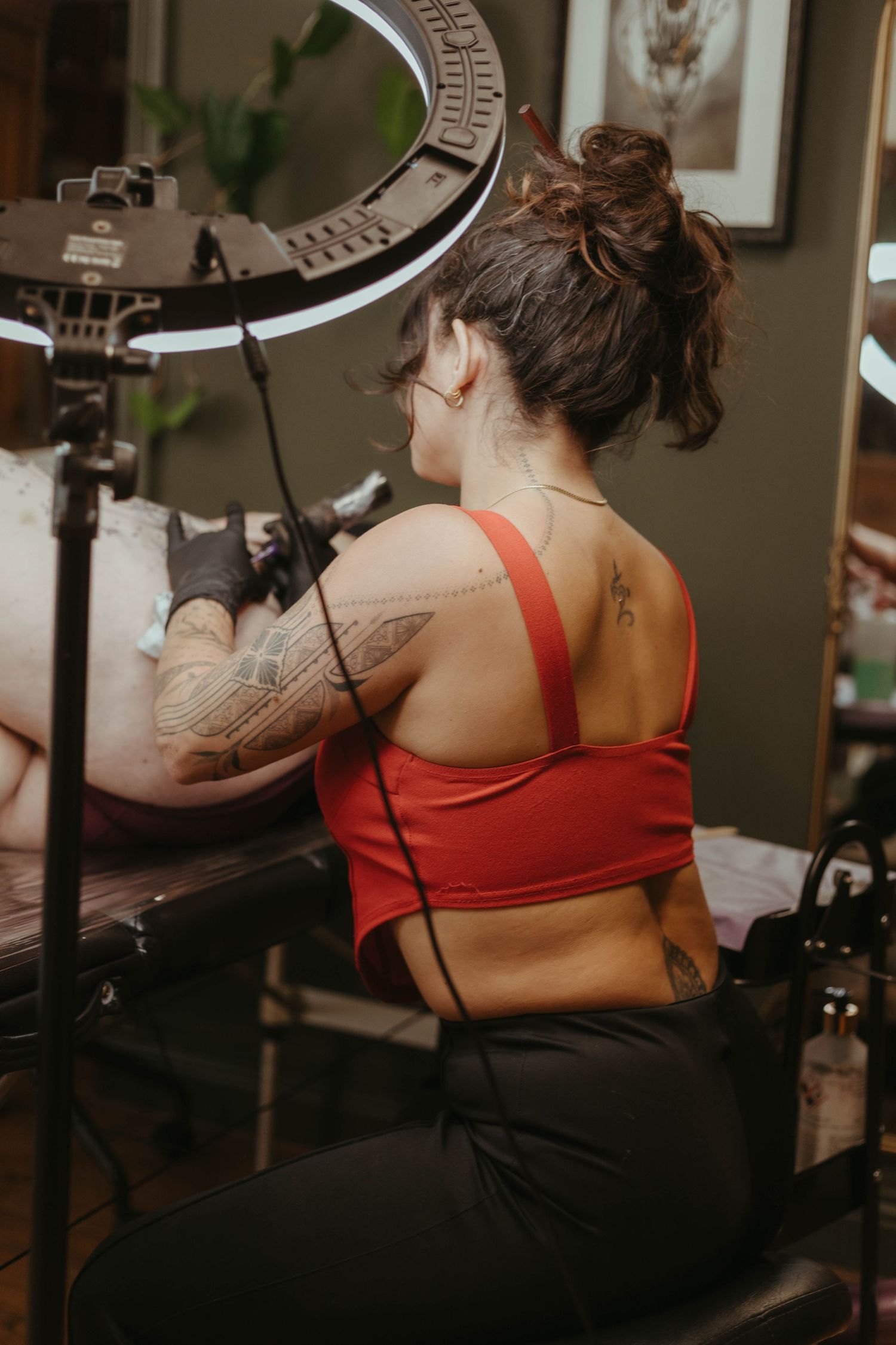 Photo of tattoo artist tattooing a client, she has her hair tied up and is wearing a red top