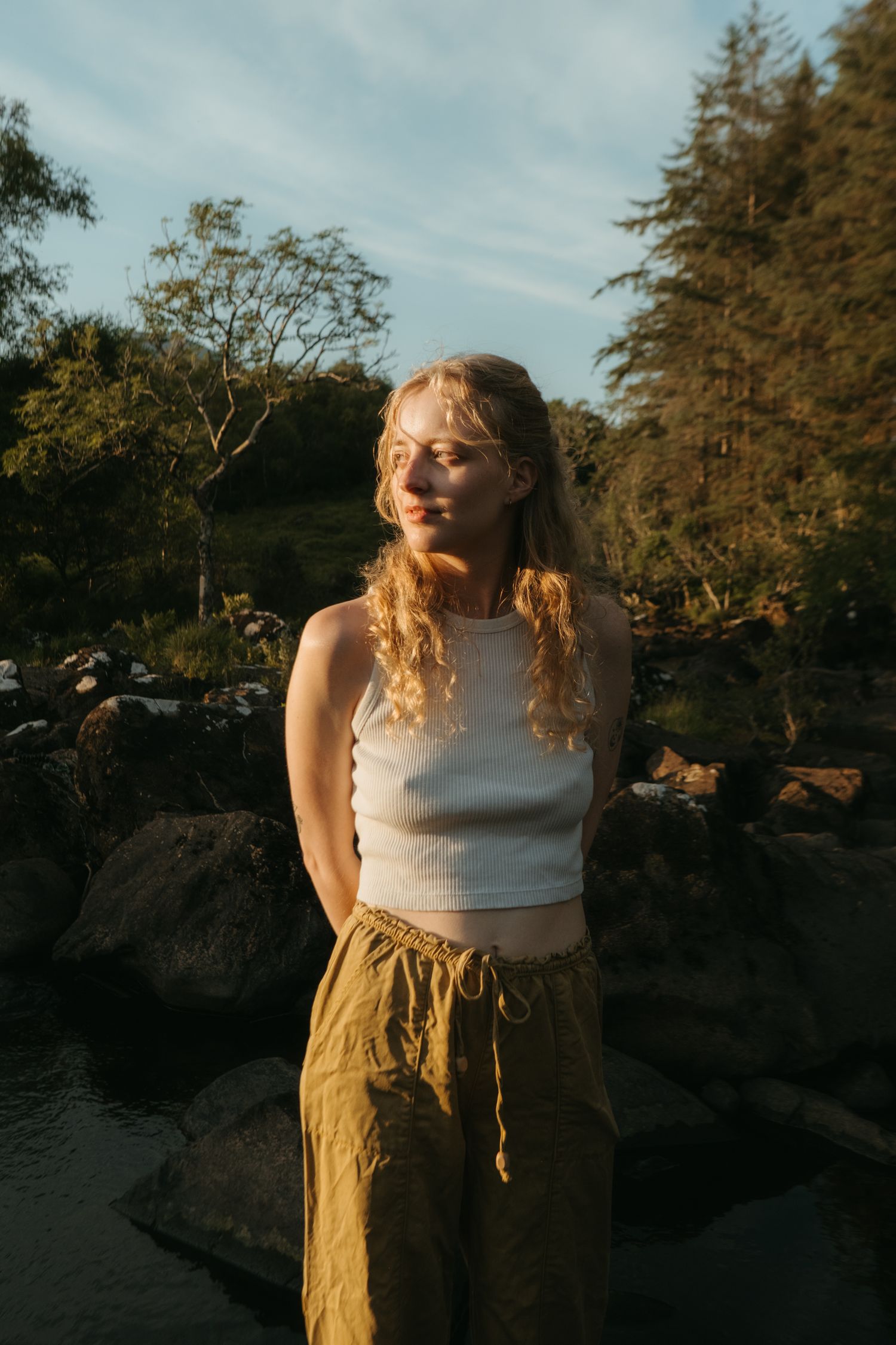 Photograph of Jacky, taken in front of a creek surrounded by rocks and trees, wearing a white top and yellow pants