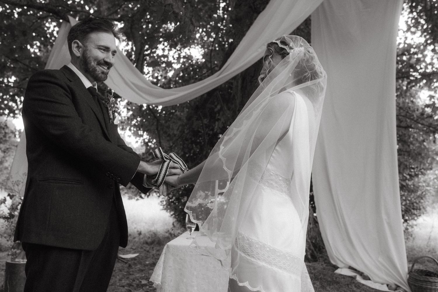 Black and white photograph of bride and groom looking at each other, during the traditional hand fasting, wind is blowing the brides veil.