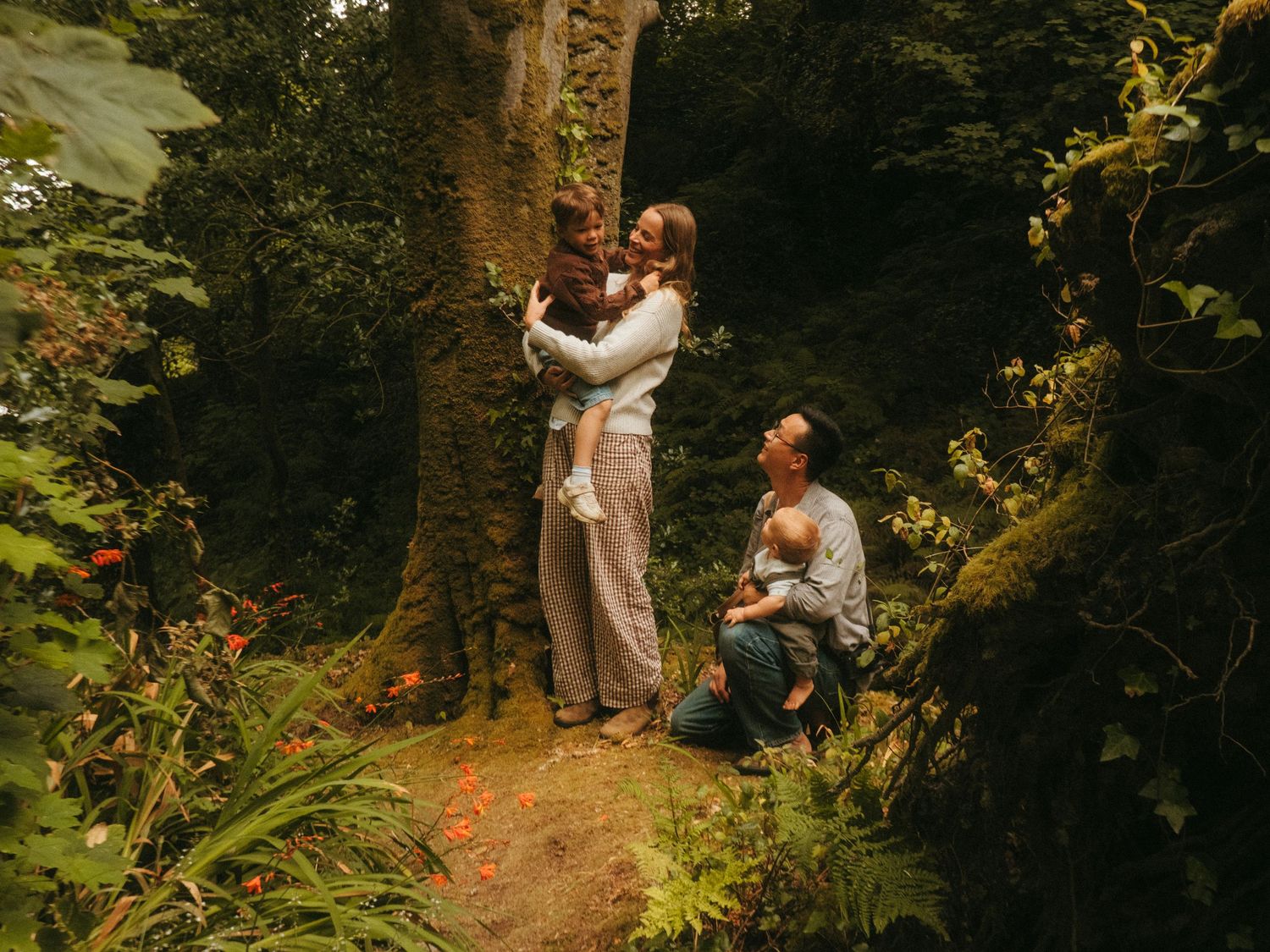 Photograph from a family shoot, you can see both parents hold on of their sons next to a lush tree, the photo is taken through leafs