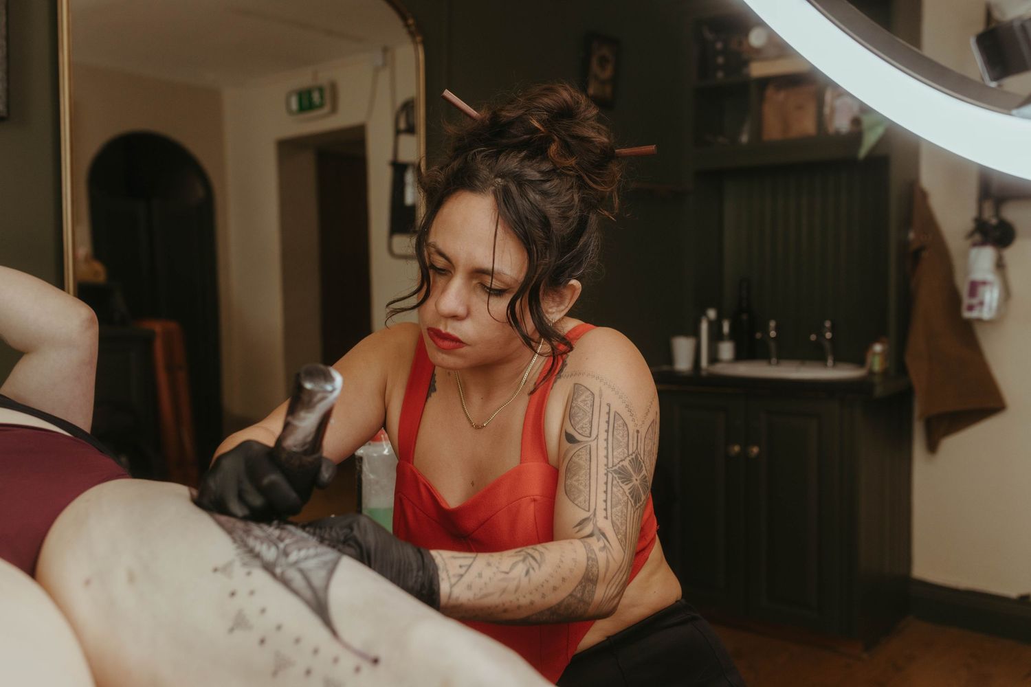 Photo of tattoo artist tattooing a client, she has her hair tied up and is wearing a red top