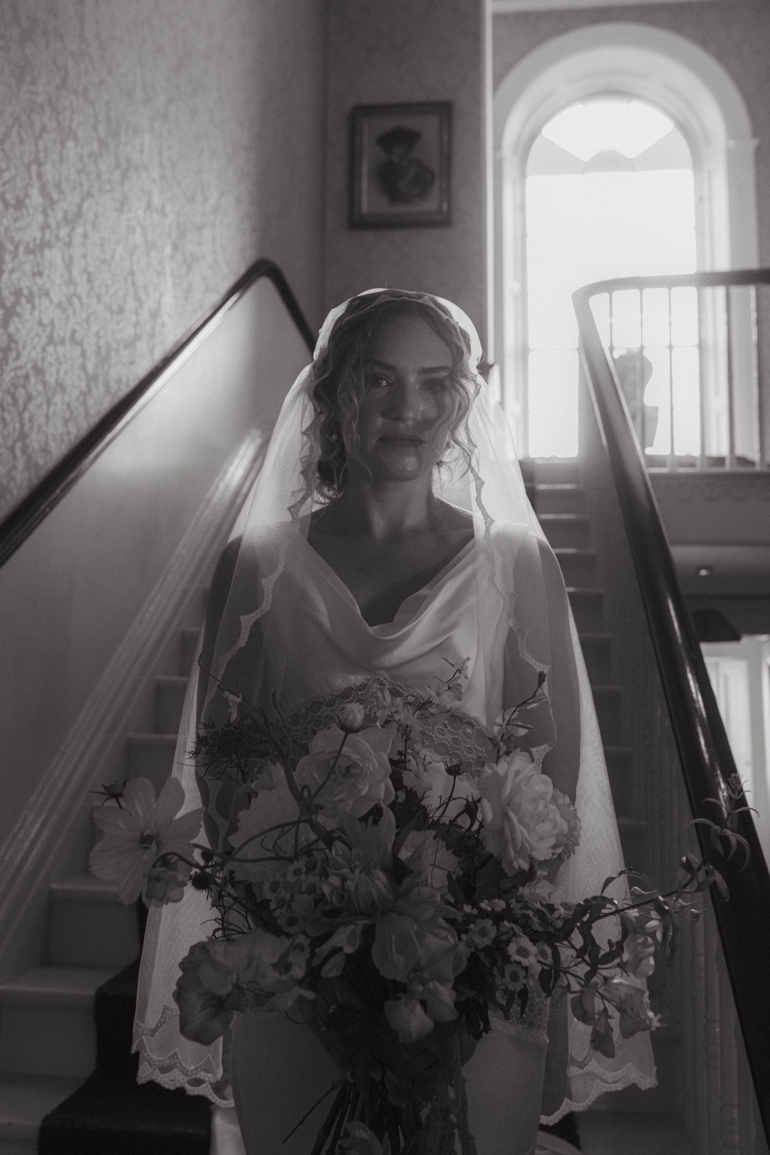Black and white photo of bride walking down the stairs at Whitestown House in Dublin. She's holding her bouquet, backlit, soft shadows.